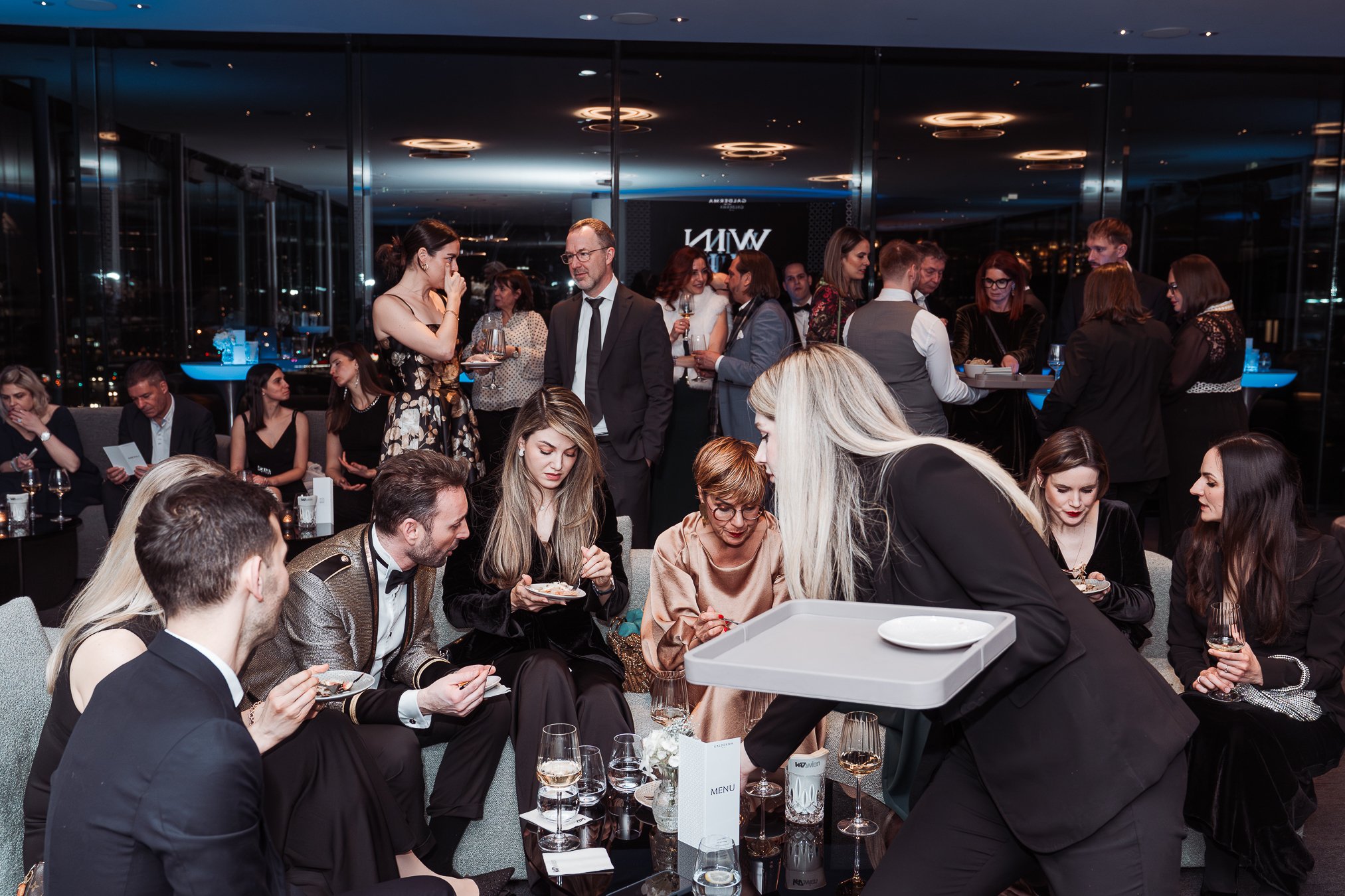 Event photography Vienna.People socializing at a formal event in a modern indoor setting, some seated and some standing, with drinks and food on the tables.