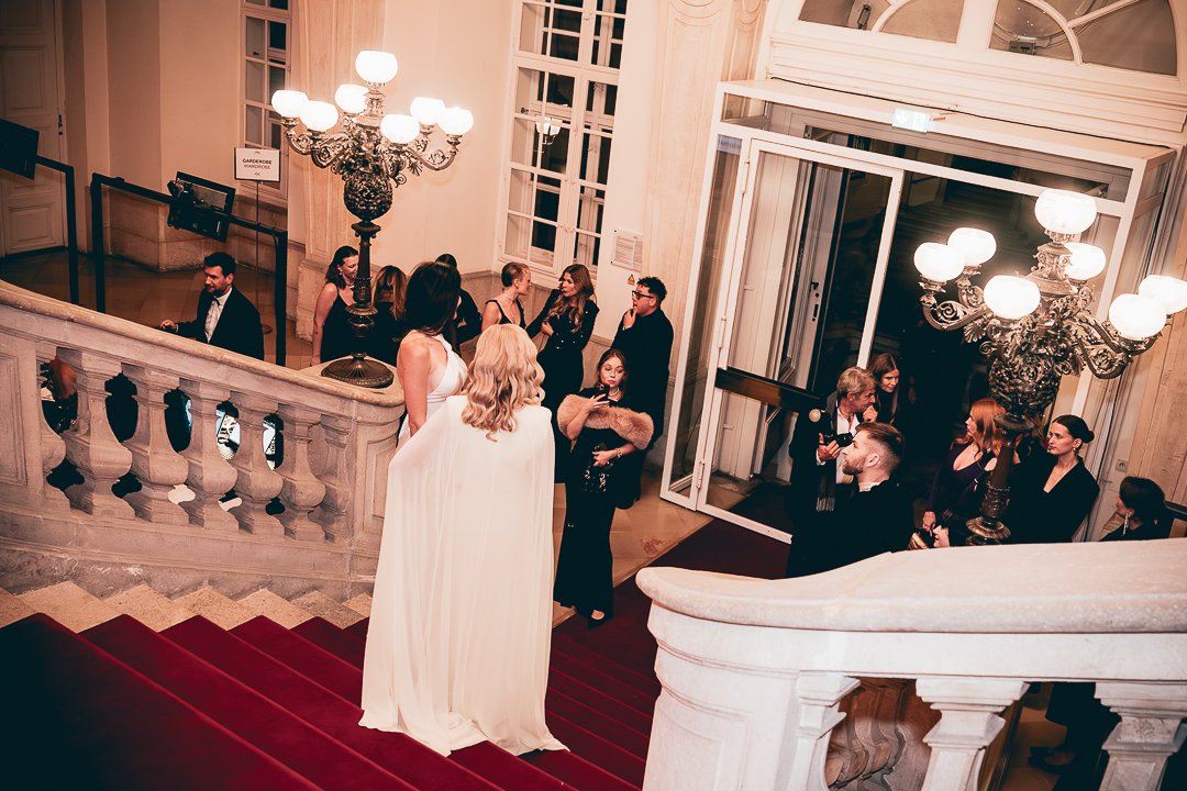 Vienna Awards!People gathering inside a grand, elegant building with large chandeliers, high ceilings, and a staircase with a red carpet. Some people are taking photos while others are engaged in conversation.