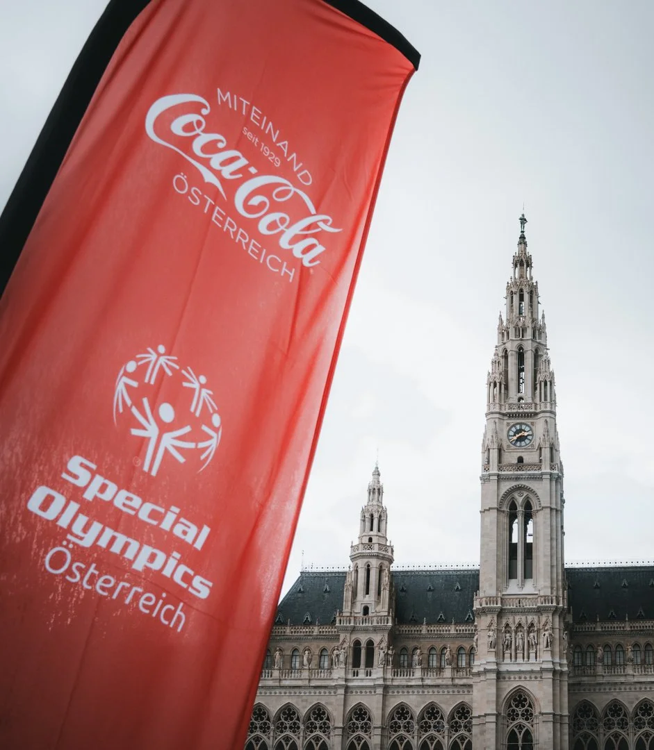 A tall red banner with the Coca-Cola logo and text promoting Special Olympics Austria, positioned in front of a historic building with Gothic architecture and a clock tower.