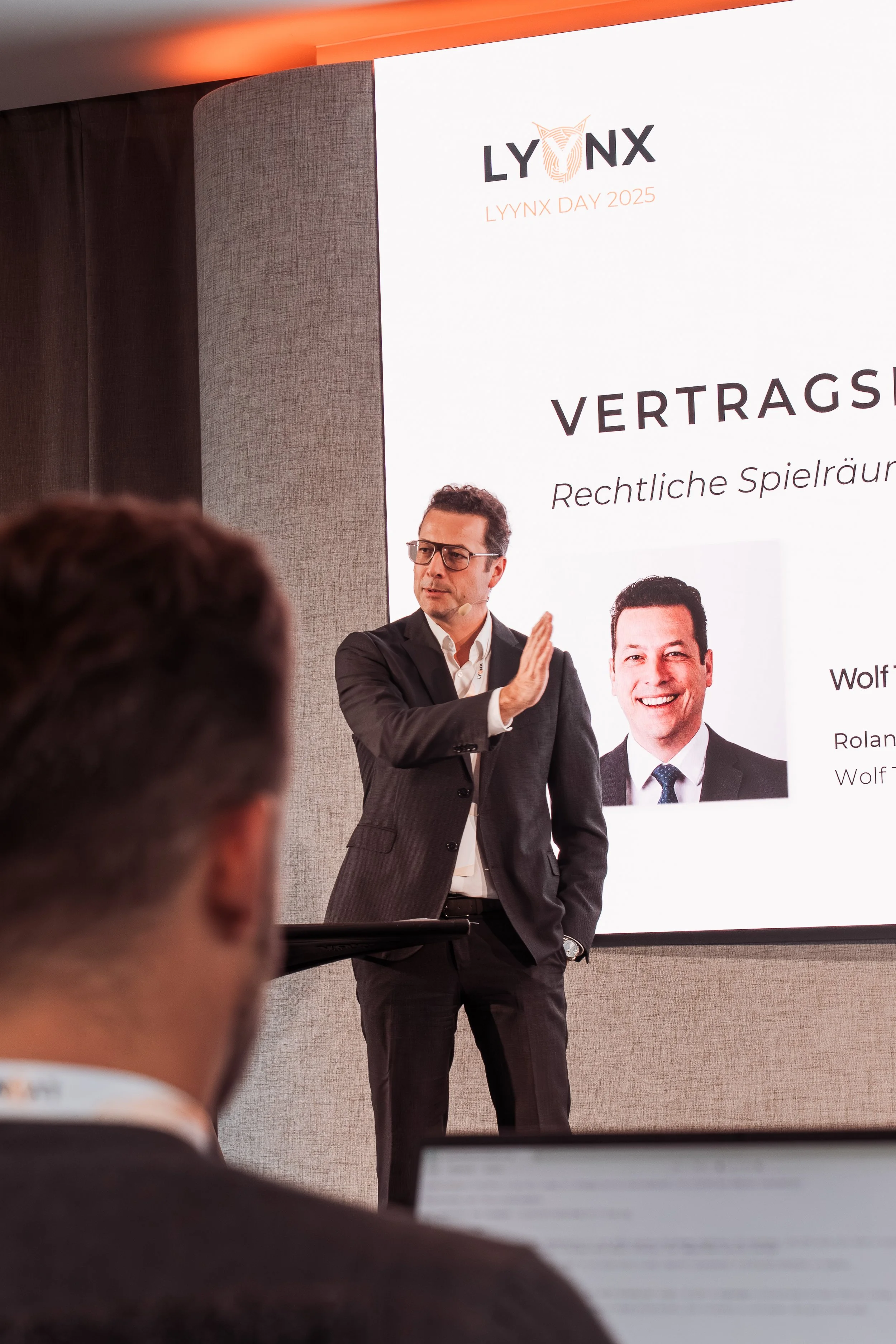 Lyynx event A man in a suit and glasses giving a presentation at an event, with a large screen behind him displaying the text 'LYNX Day 2025' and a headshot of another man. The audience is visible, with people taking notes.
