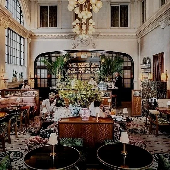 Elegant restaurant interior with high ceilings, large windows, chandeliers, and vintage decor. Several groups of people are dining, surrounded by lush plants and stylish furniture.