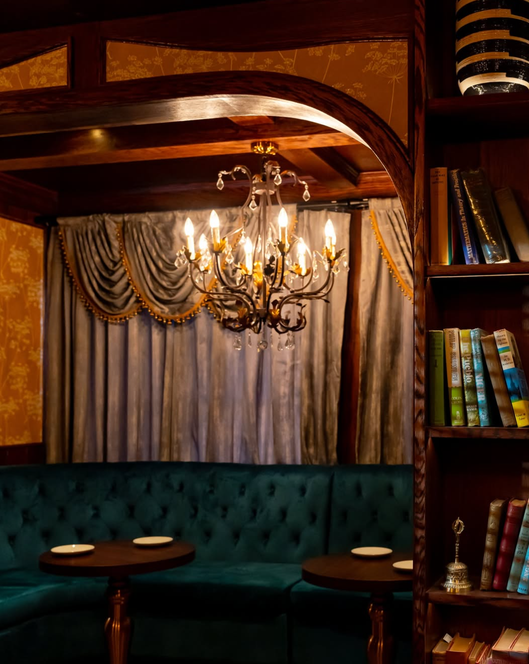 An elegant room with a chandelier, thick curtains, a curved green velvet sofa, and a wooden bookshelf filled with books.