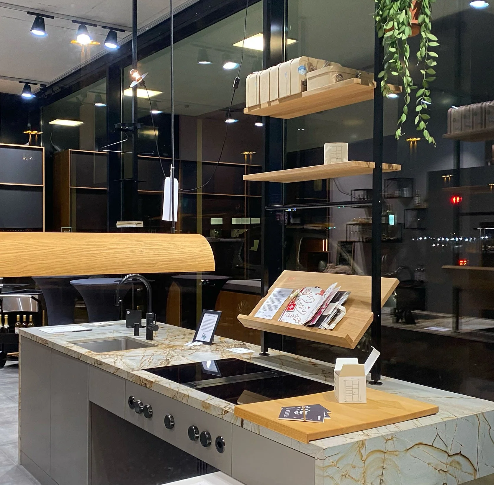 Moderne Kücheneinrichtung mit Küchenarbeitsplatte aus Marmor, integriertem Herd, schwarzer Spüle und schwarzen Küchenarmatur, umgeben von Holzregalen mit Geschirr und Dekor. Im Hintergrund große Fensterscheiben und Restaurantbeleuchtung bei Nacht.