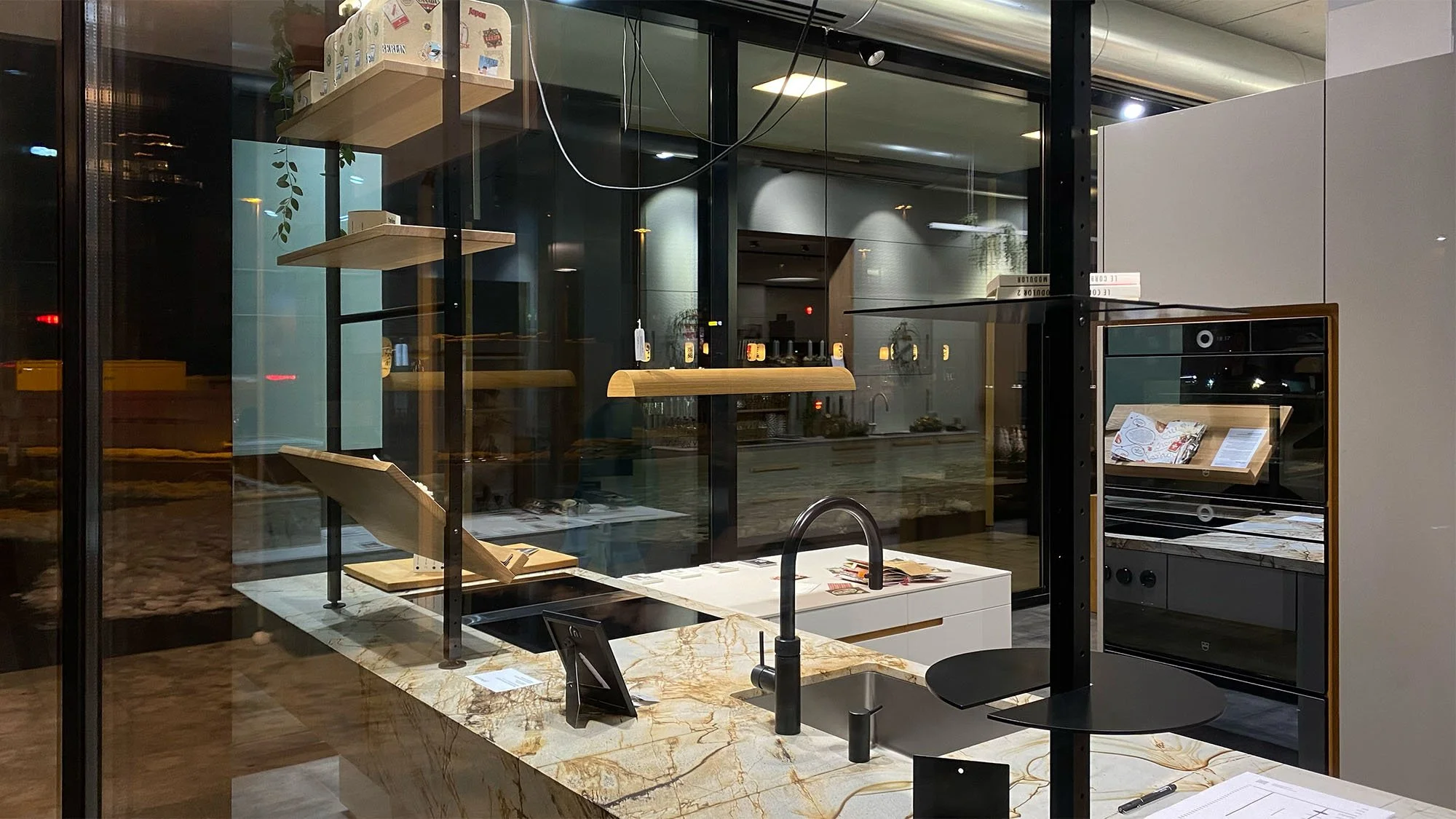 Moderne Küchenausstellung mit Marmorarbeitsfläche, schwarzen Wasserhahn, eingebauten Backofen und Mikrowelle, offene Regale mit Geschirr und Dekoration, großes Fenster, reflektierende Glasfronten