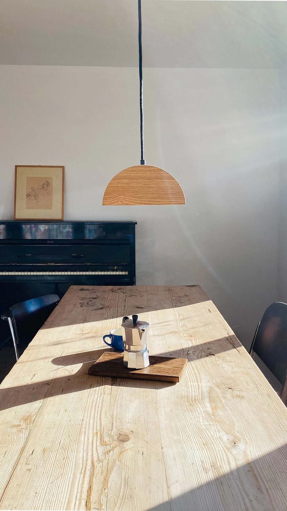 Ein Holztisch mit einer Moka-Kanne, einer blauen Kaffeetasse und einem Holzbrett, Sonnenlicht strahlt auf den Tisch, im Hintergrund ein Klavier und ein Bild an der Wand, eine hängende Lampe aus Holz über dem Tisch.