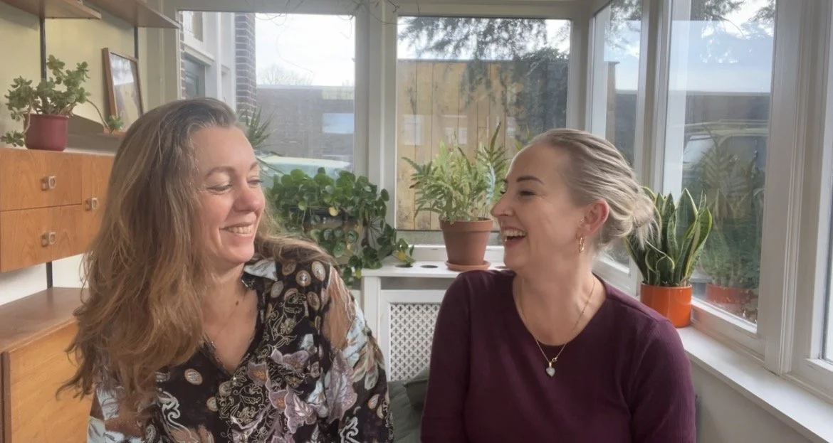 Twee vrouwen lachen naar elkaar in een kamer met veel planten en grote ramen.