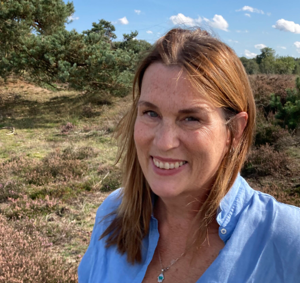 Een vrouw met lang, lichtbruin haar in een blauwe blouse staat buiten, met een landschap van groen en paarse heide op de achtergrond.