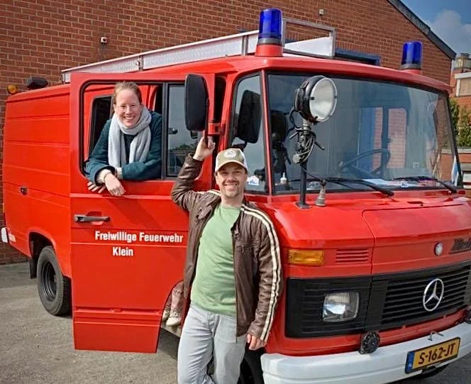 Twee mensen poseren voor een rood brandweervoertuig met blauwe zwaailichten, waarop de tekst 'Freiwillige Feuerwehr Klein' staat.