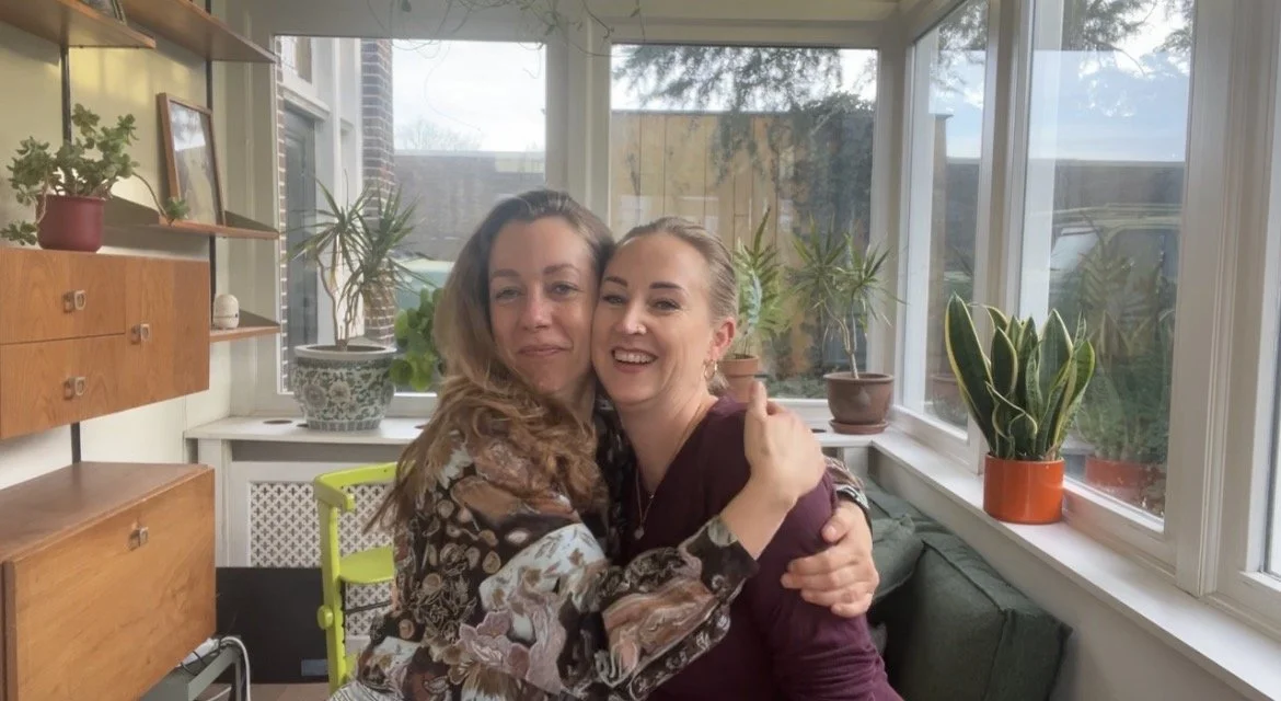 Twee vrouwen omhelzen elkaar vrolijk in een zonnige serre met planten op de vensterbanken.