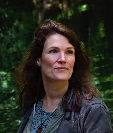 Portret van een vrouw met bruin haar, outdoors met groene bomen op de achtergrond.