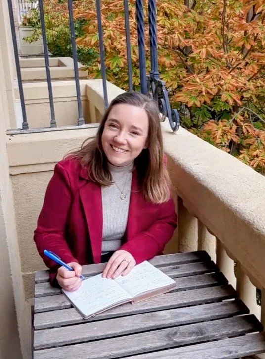 Sabrina sitting at a table on a balcony writing in a notebook
