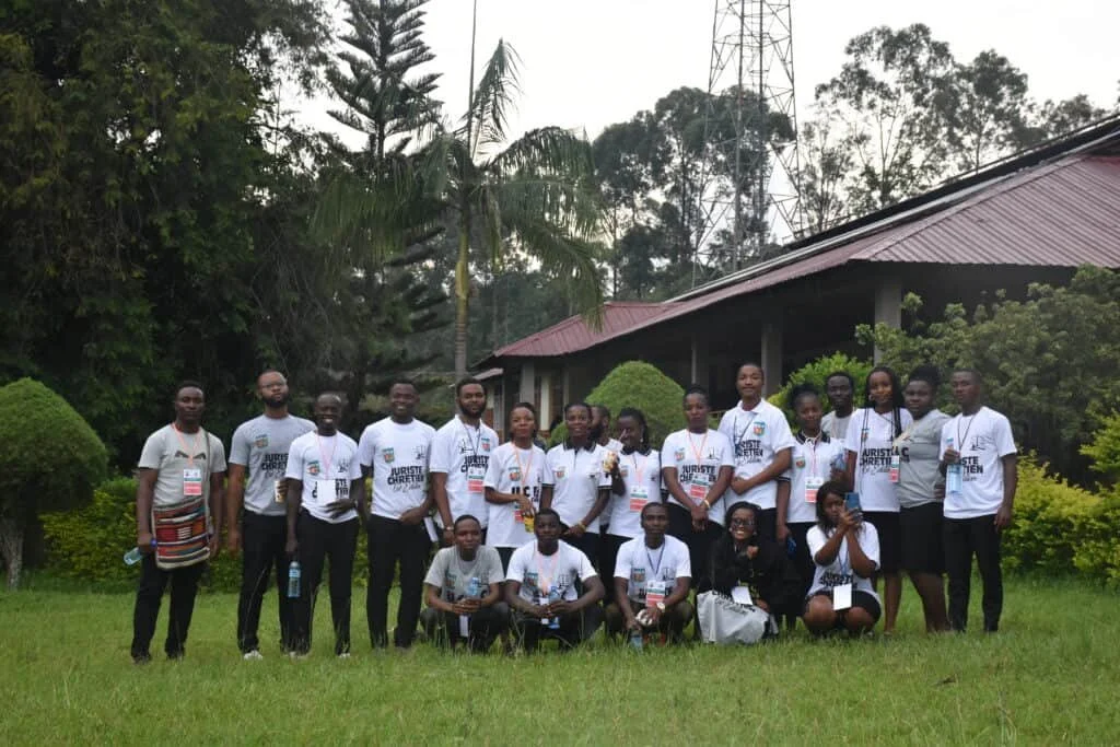 A group of young students standing on a grassy area outdoors, wearing matching white t-shirts with a logo and text, with trees and a building with a red roof in the background.