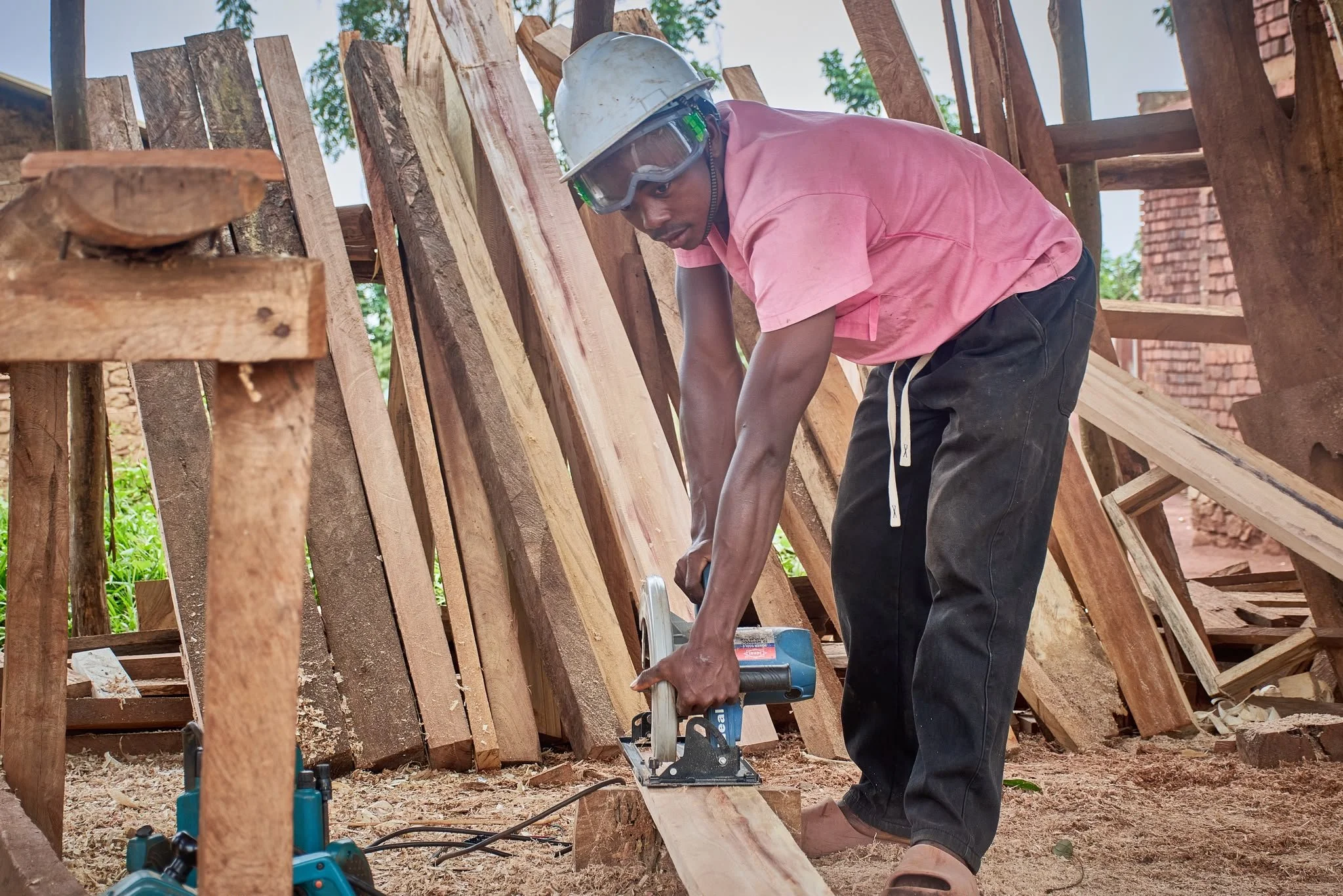 A man in pink shirt, safety glasses, and a white helmet working on a woodworking project outdoors with wooden planks and tools around him.