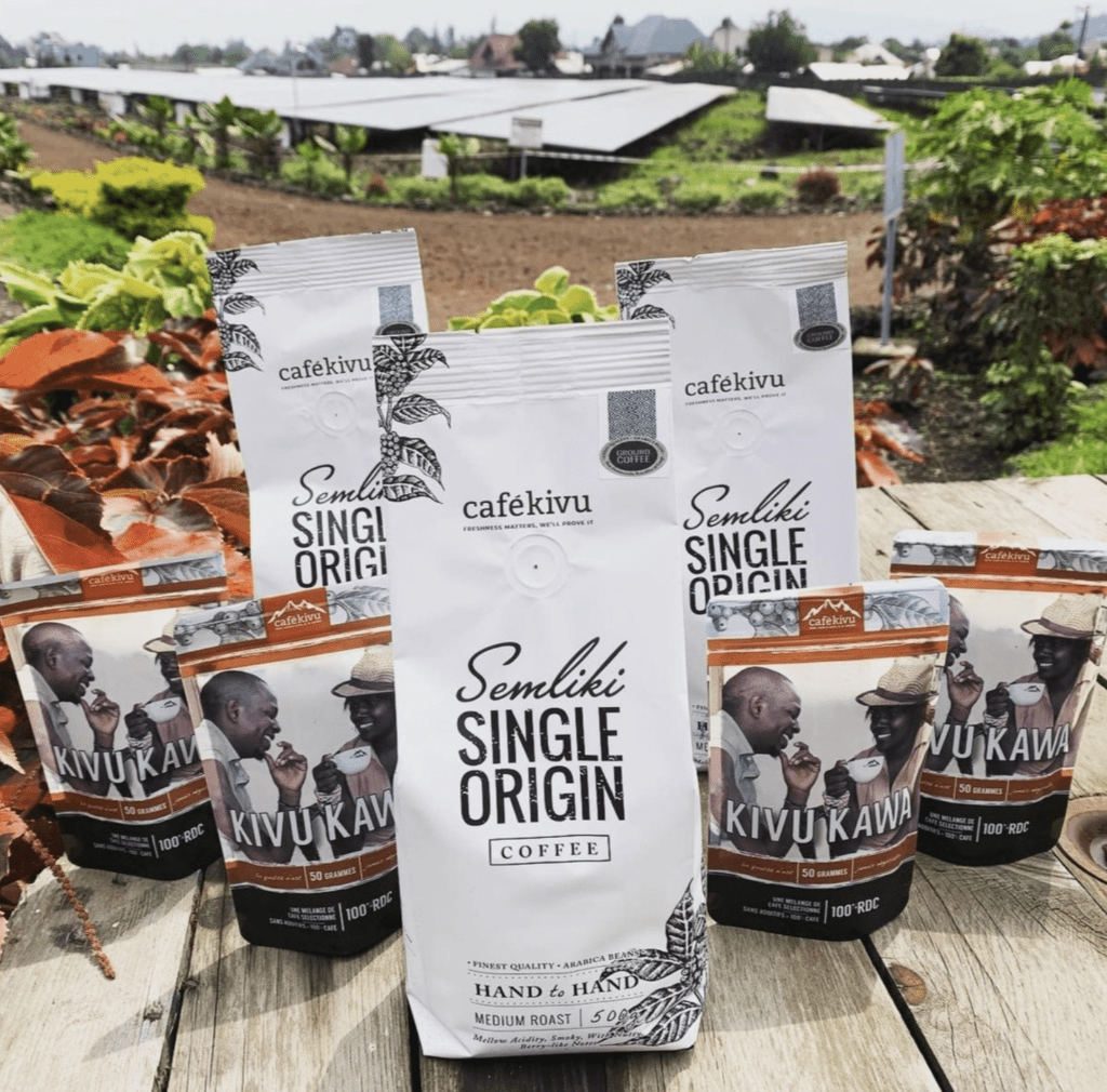 Several packages of coffee labeled 'Semliki Single Origin' and 'Kivu Kawa' placed on a wooden surface outdoors, with green plants and solar panels in the background.