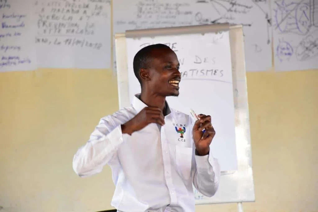 A happy man is standing and speaking in front of a whiteboard, holding a pen in his hand. He is wearing a white shirt with a logo on the chest. The background includes written text and diagrams on the whiteboard and walls.