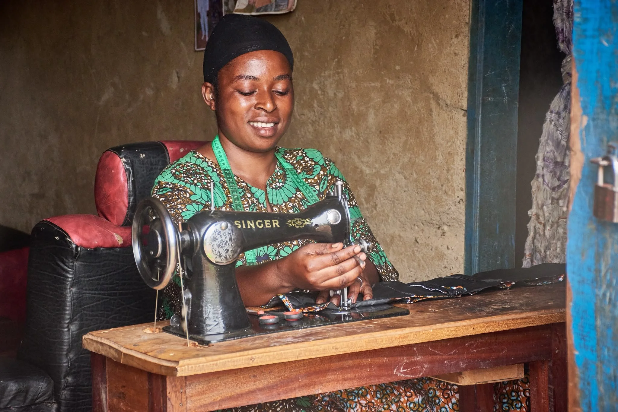 A woman sitting at a wooden table with a sewing machine, wearing a batik dress, a black head covering, and a measuring tape around her neck. She is smiling and looking down at the sewing machine.