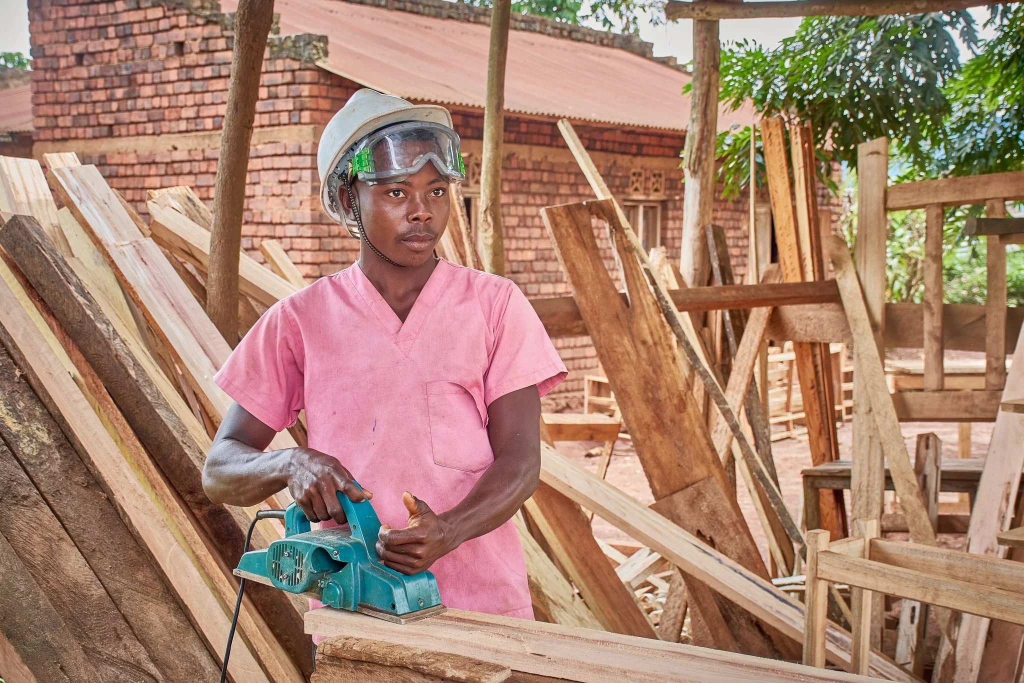 A person wearing a pink shirt and safety gear using a power sander on a wooden piece at a construction site with wood framing and a brick building in the background.