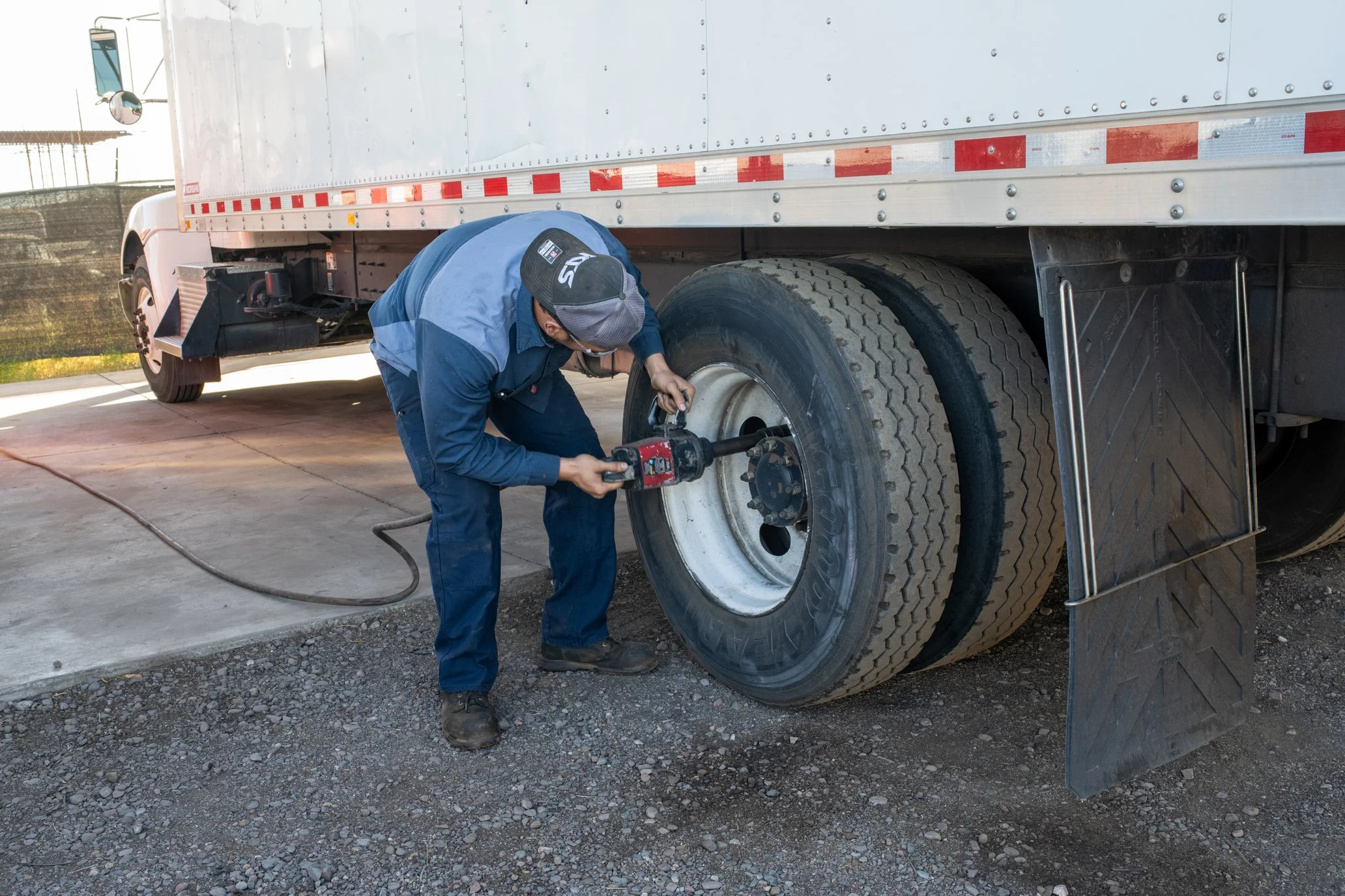 A mechanic working on the front wheel of a large truck, using a power tool, with a concrete and gravel surface and part of the truck trailer visible.