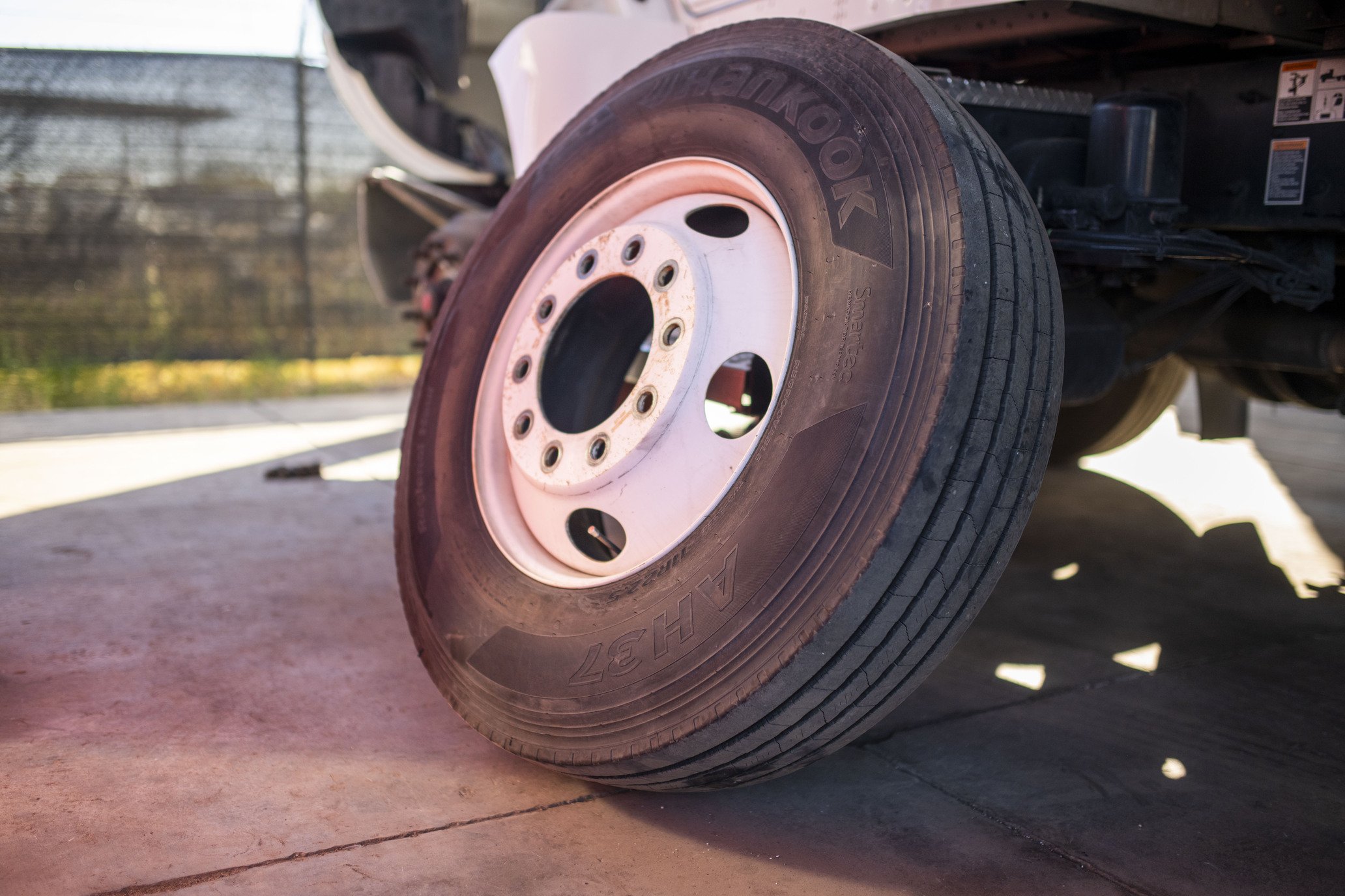 Close-up of a truck tire with a white rim on a concrete surface with a fence and trees in the background.