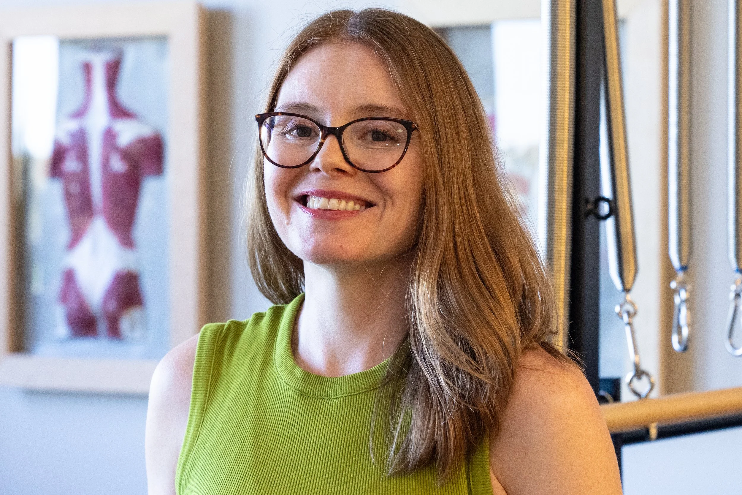 A smiling woman with long red hair and glasses wearing a green sleeveless top, in a fitness or physical therapy setting with anatomical poster and exercise equipment in the background.