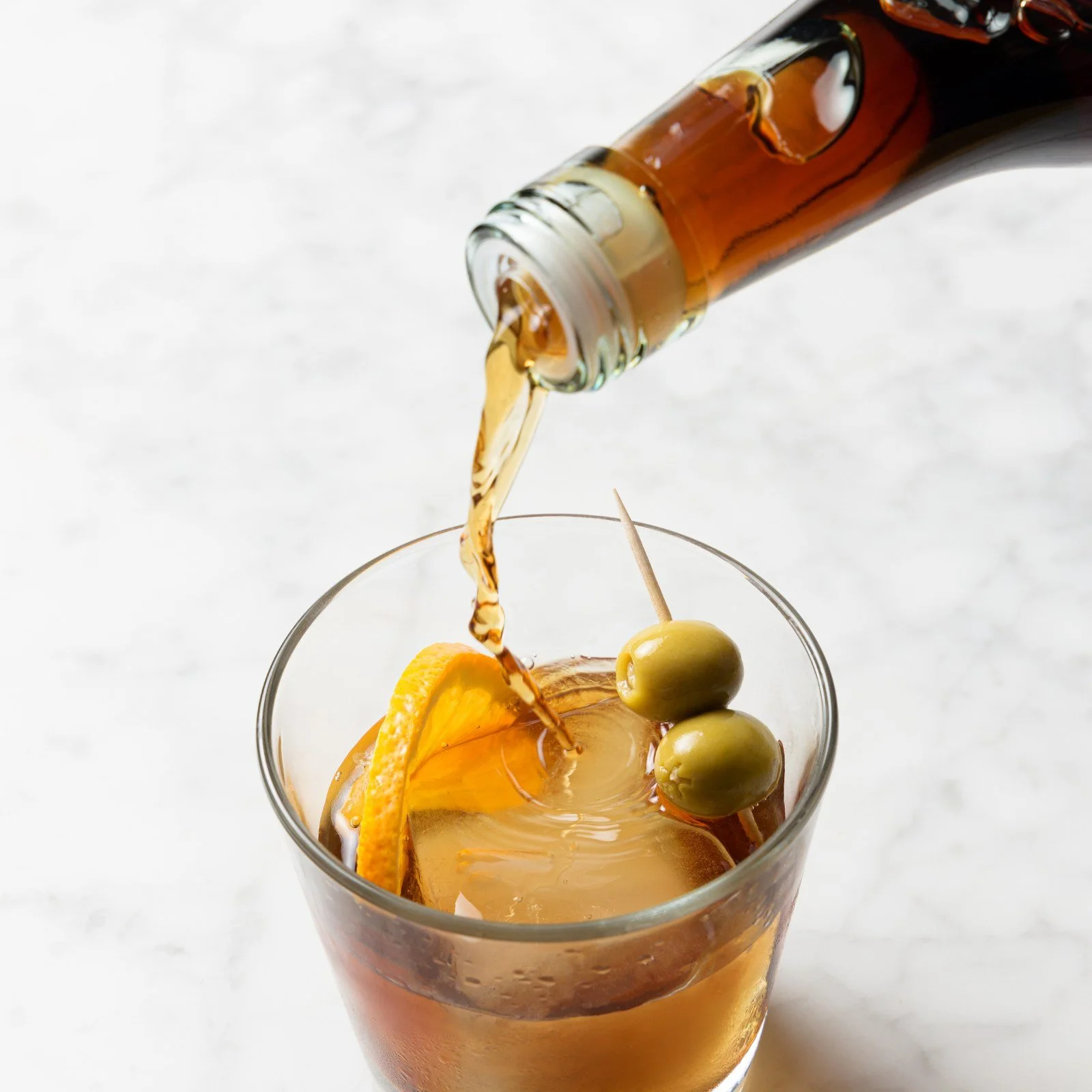 Vermouth being poured into a short glass with ice, lemon slice and olives.