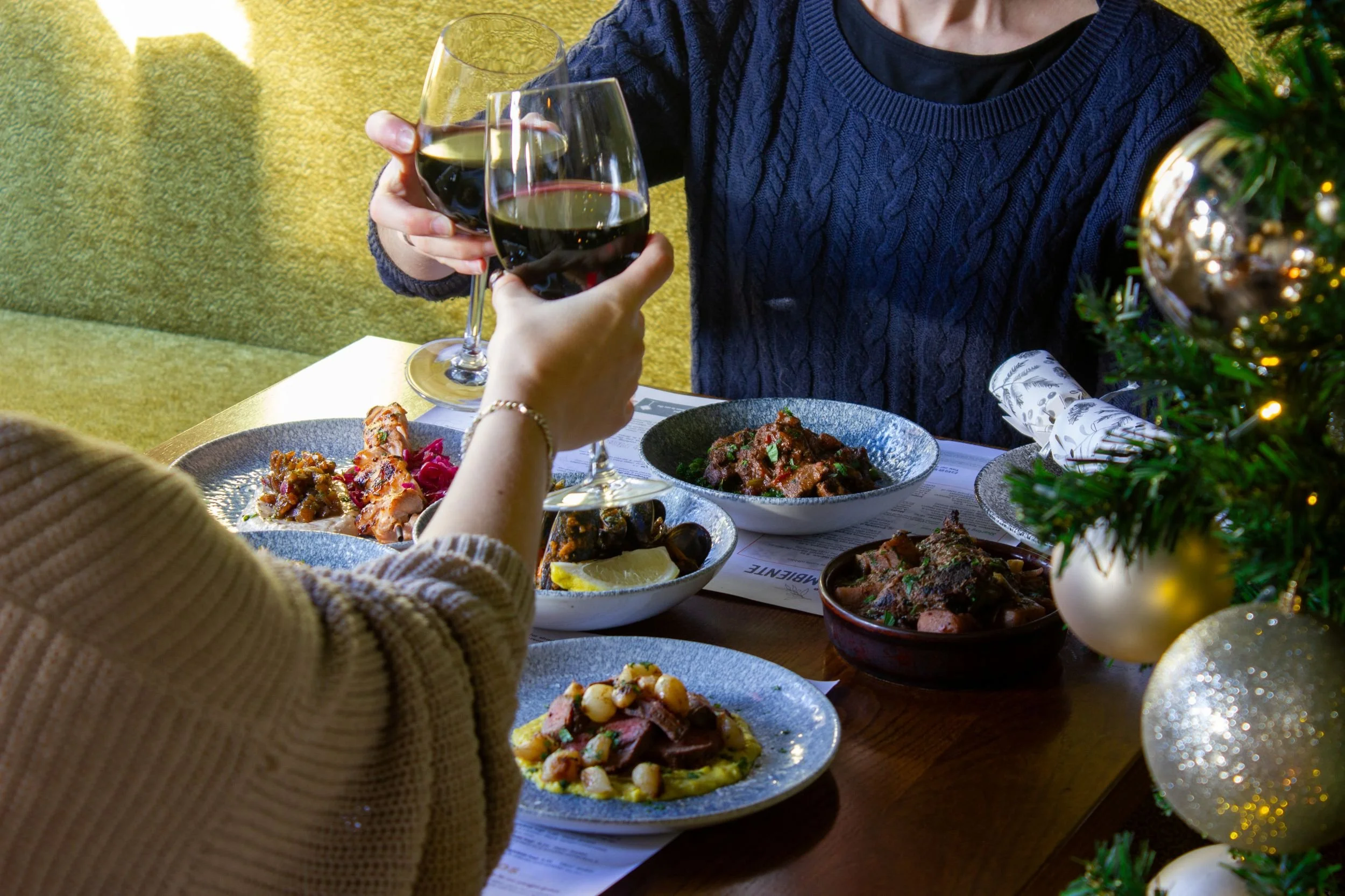 Two people cheersing with a glass of wine over tapas with Christmas tree in background