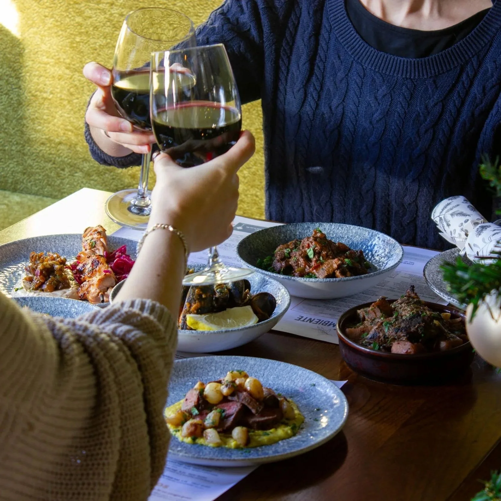 Two people cheersing with glasses over a table of festive tapas