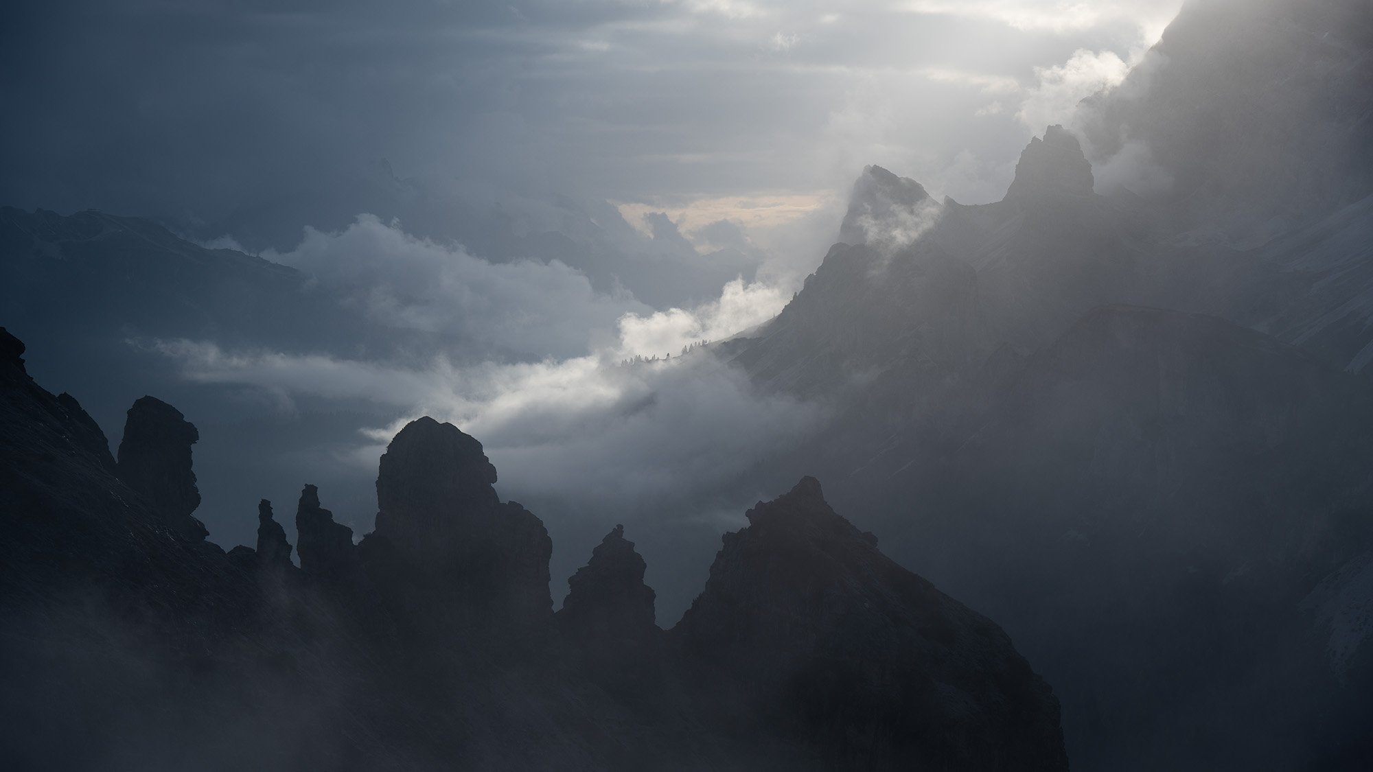 Mountain range with jagged peaks shrouded in mist and clouds, with sunlight breaking through in the background.