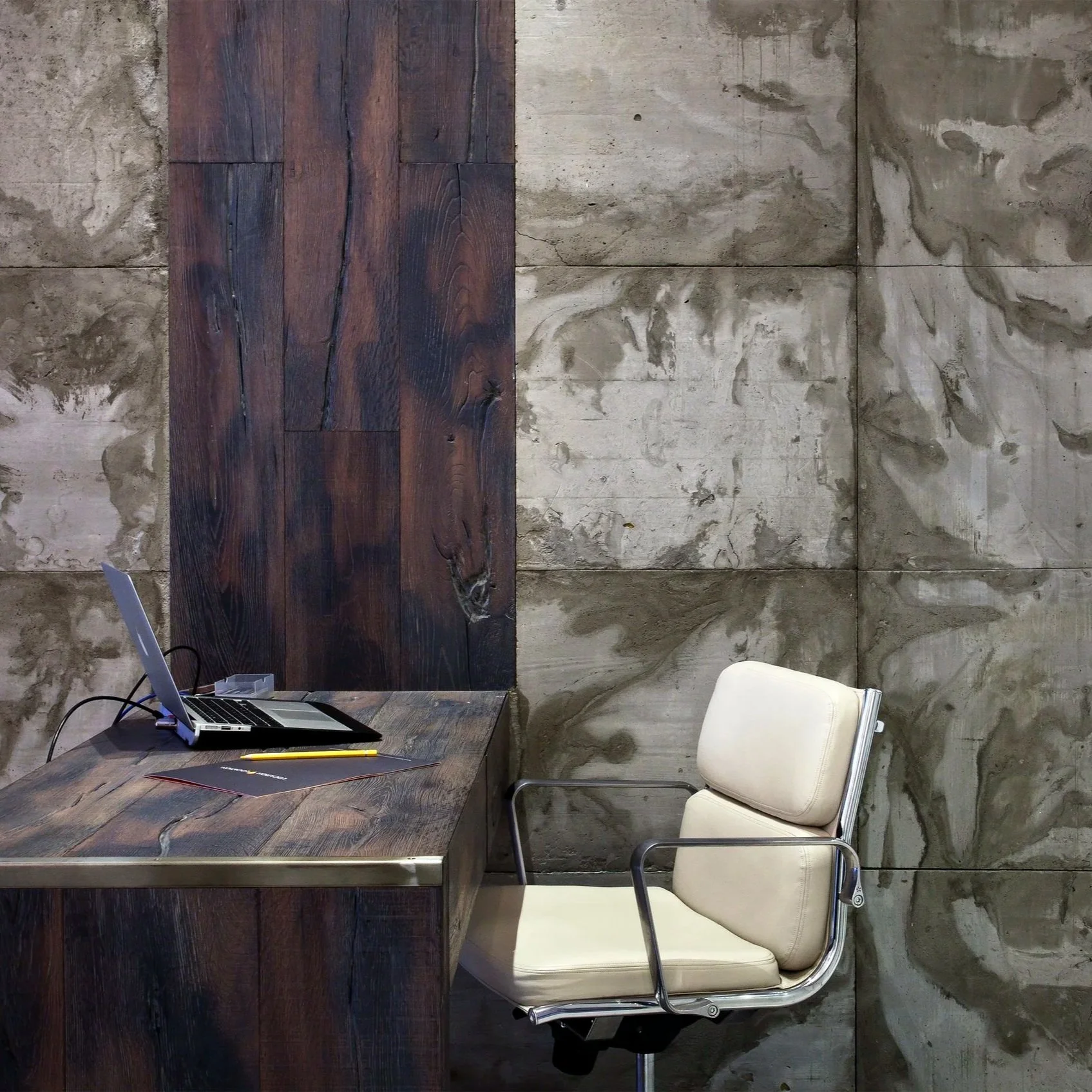 Empty office space with a wooden desk, a beige office chair, a laptop, and a notepad with a pen, against a textured concrete wall with a wooden panel.
