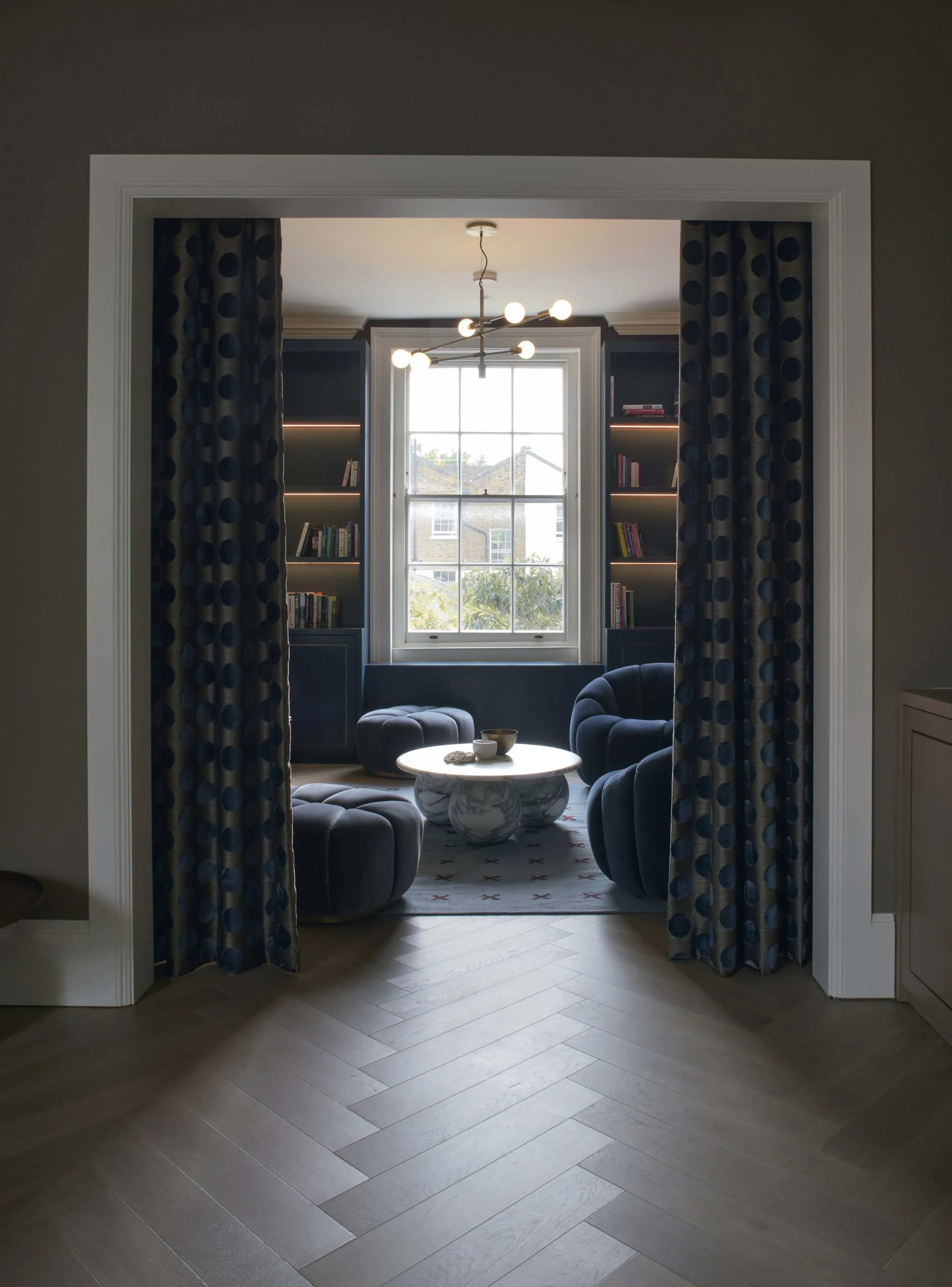 Navy library snug in Notting Hill with built-in bookshelves, curved velvet armchairs, marble coffee table and herringbone oak flooring, framed by patterned curtains