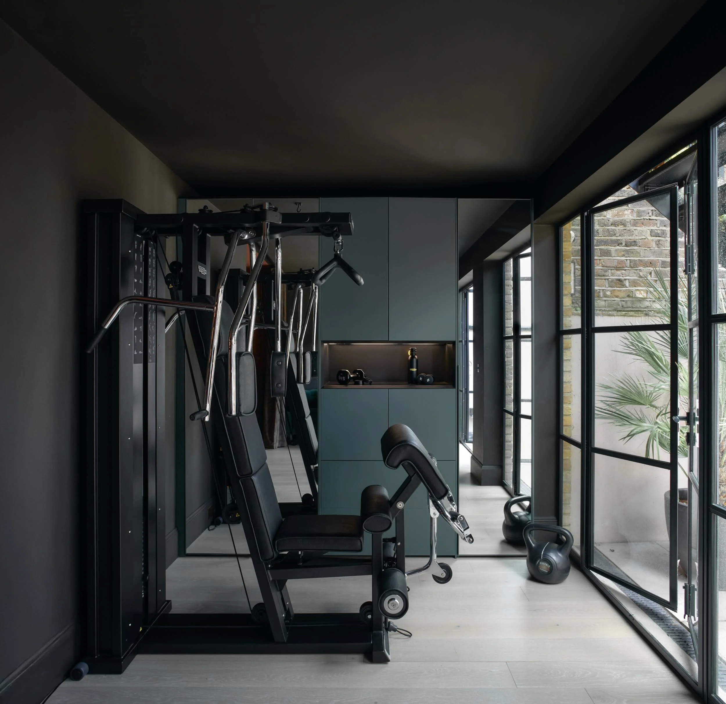 Home gym in Notting Hill with dark green cabinetry, multi-gym equipment, kettlebells and Crittall-style doors opening onto a garden