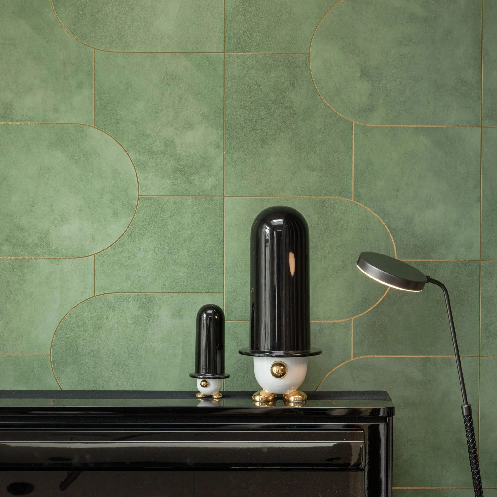 Decorative black and gold sculptures resembling rockets on a black surface against a green patterned wall, with a black floor lamp on the right.