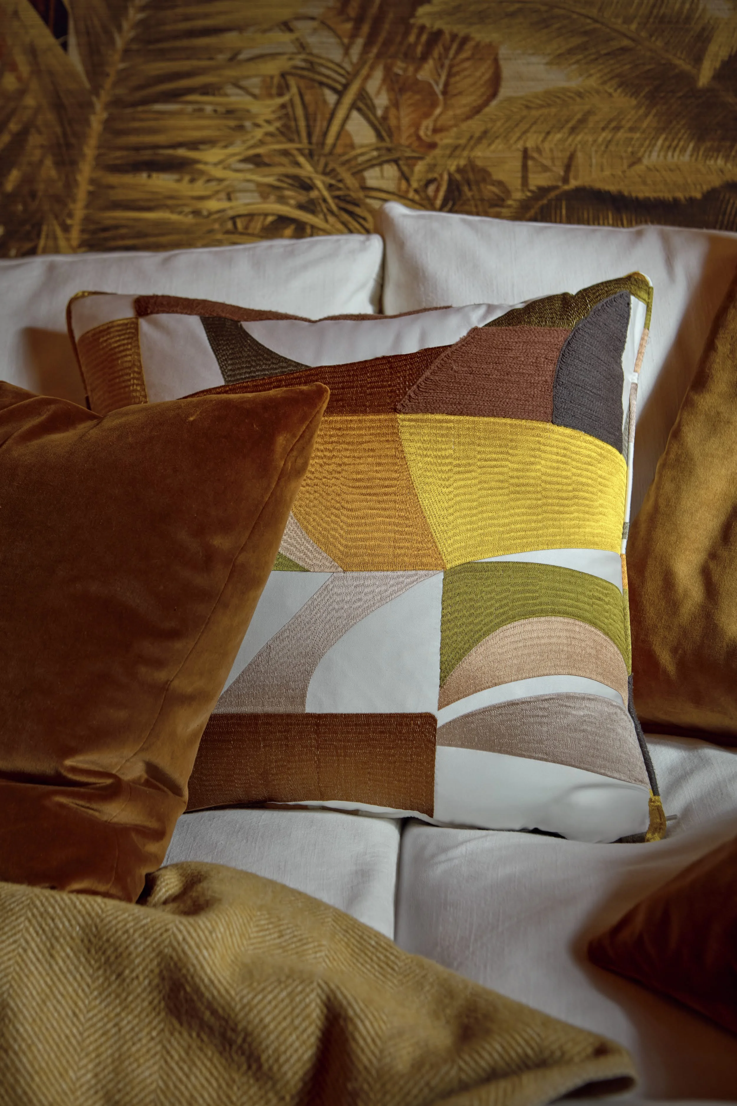 Close-up of geometric embroidered cushion in mustard, terracotta and olive tones styled with rust velvet cushions on a cream sofa, palm-print wallpaper behind