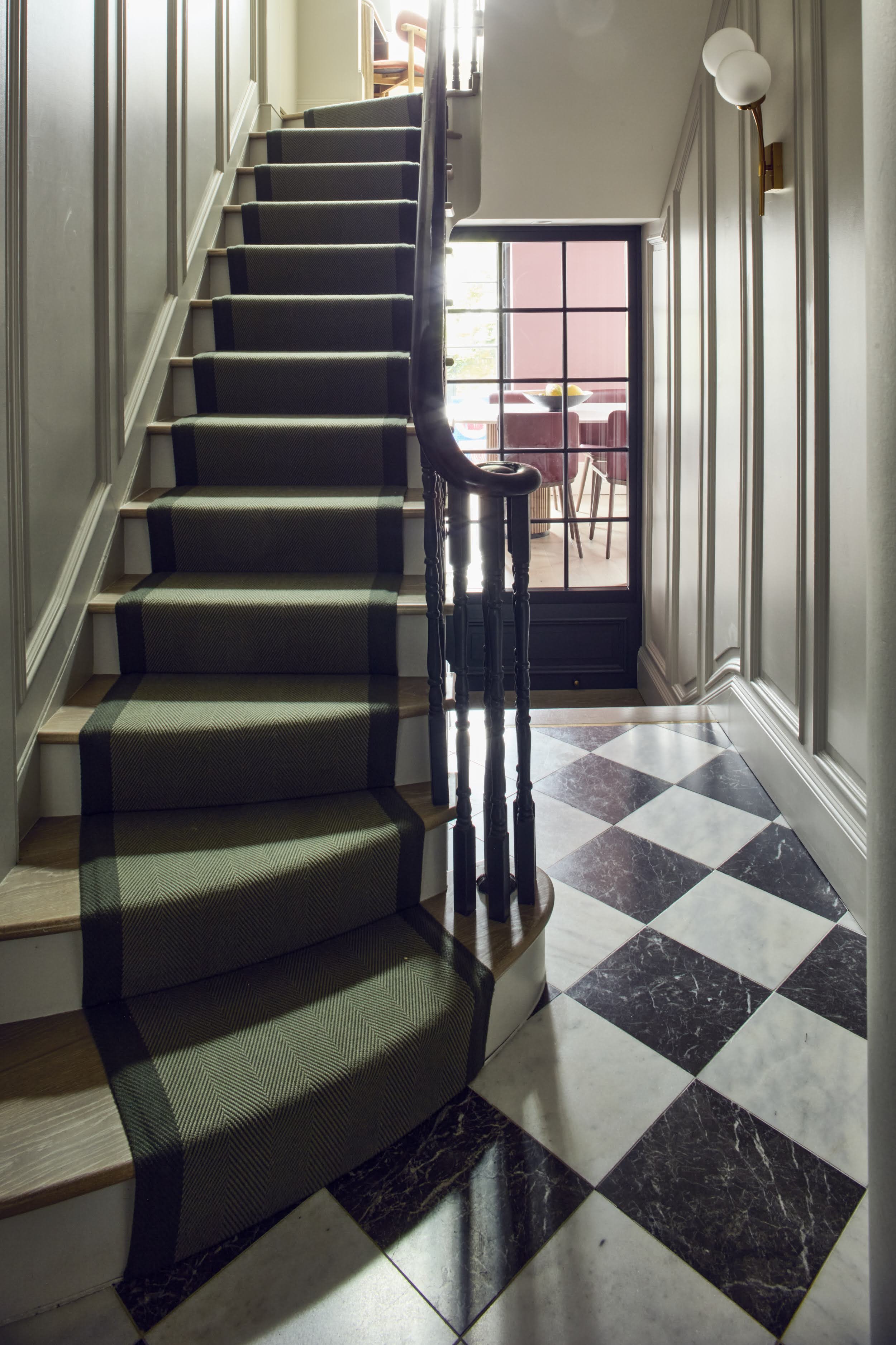Notting Hill hallway with panelled walls, green herringbone stair runner, chequerboard marble floor, and glimpse into the pink dining room beyond