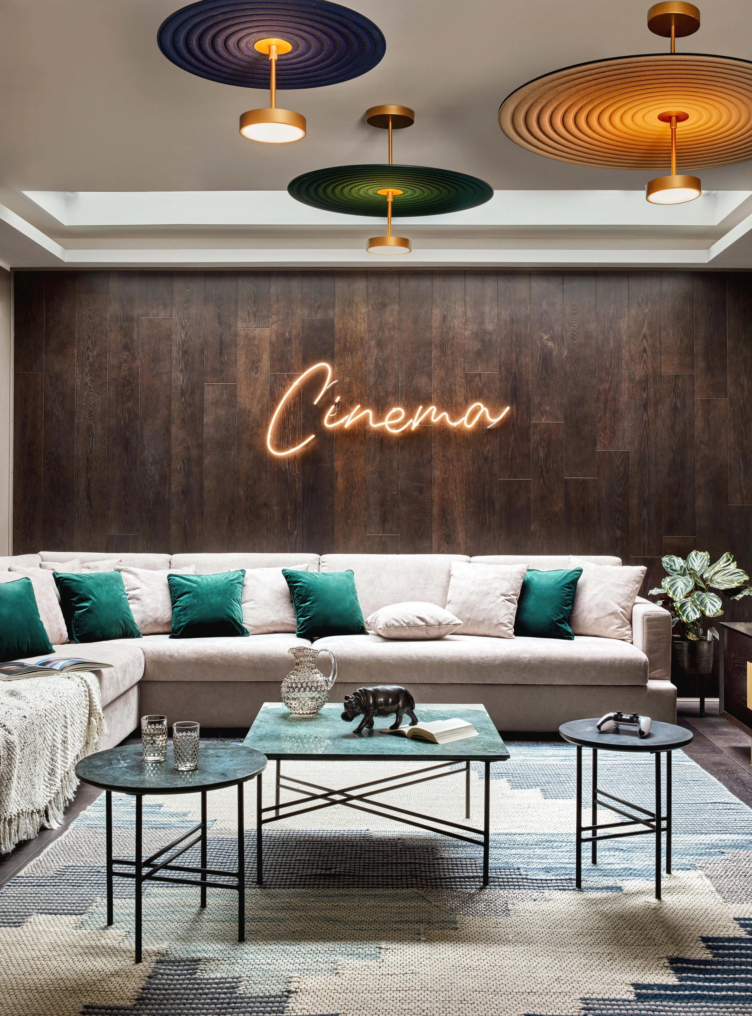 Modern living room with a beige sectional sofa with dark green pillows, a glass coffee table with decorative items, and a wooden wall with a neon sign that says 'Cinéma.' There are colorful ceiling lights and a patterned rug on the floor.