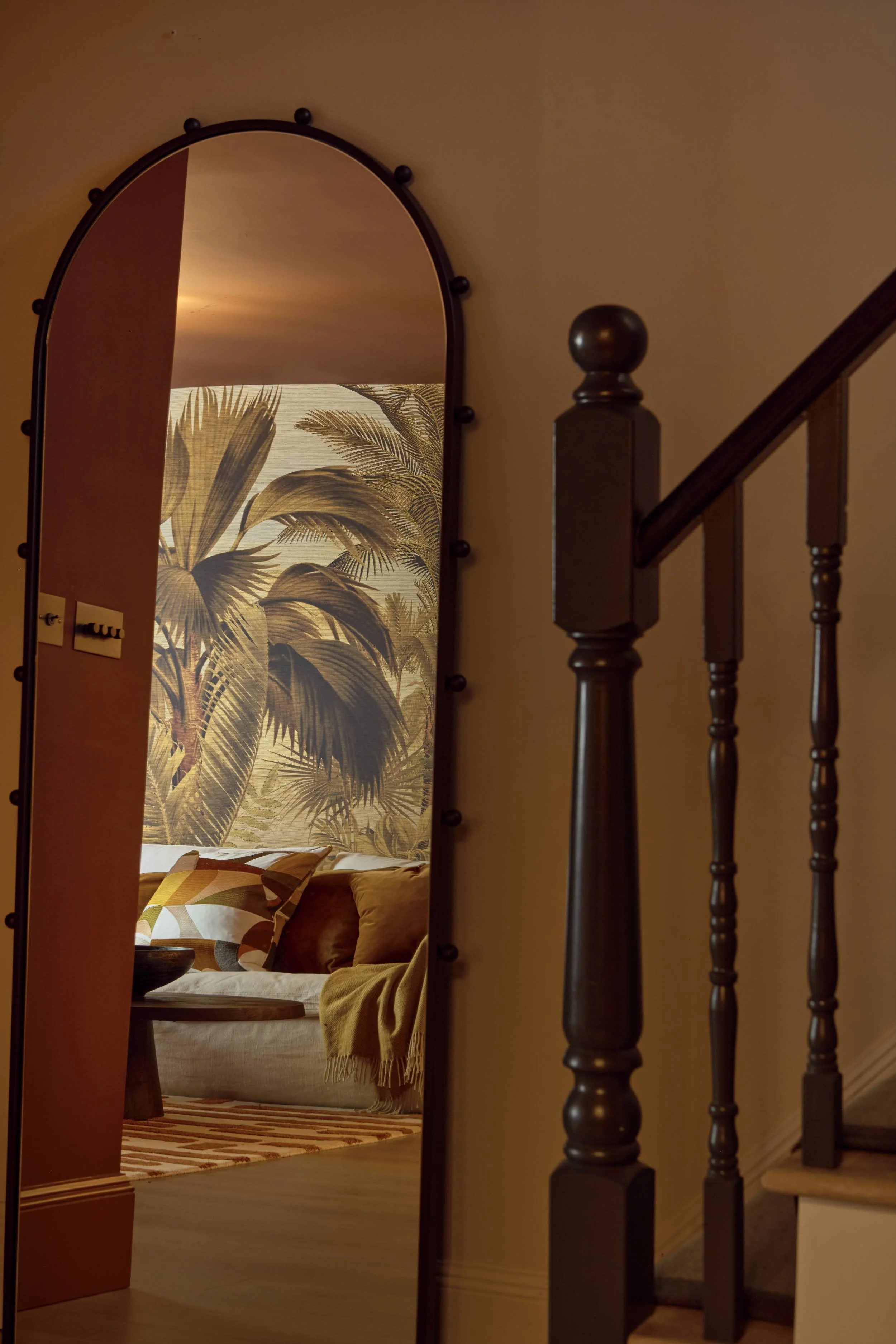 Arched black-framed mirror in a Notting Hill staircase hallway reflecting the palm-print basement cinema room, beside a traditional dark wood banister