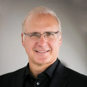Professional portrait of a middle-aged man with short gray hair and glasses, wearing a black suit and black shirt, smiling at the camera.