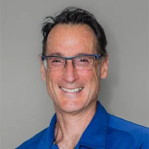 Portrait of a man with glasses, dark hair, and a friendly smile, wearing a blue shirt against a neutral background.