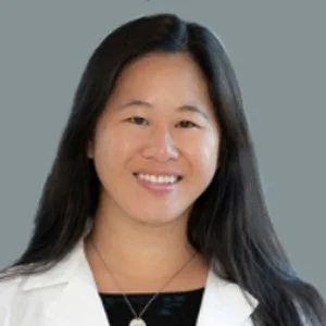 A woman with long black hair smiling at the camera, wearing a white shirt and a necklace, standing against a gray background.