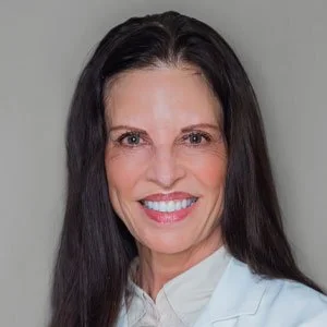 Portrait of a woman with long dark hair smiling, wearing a white shirt, against a gray background.