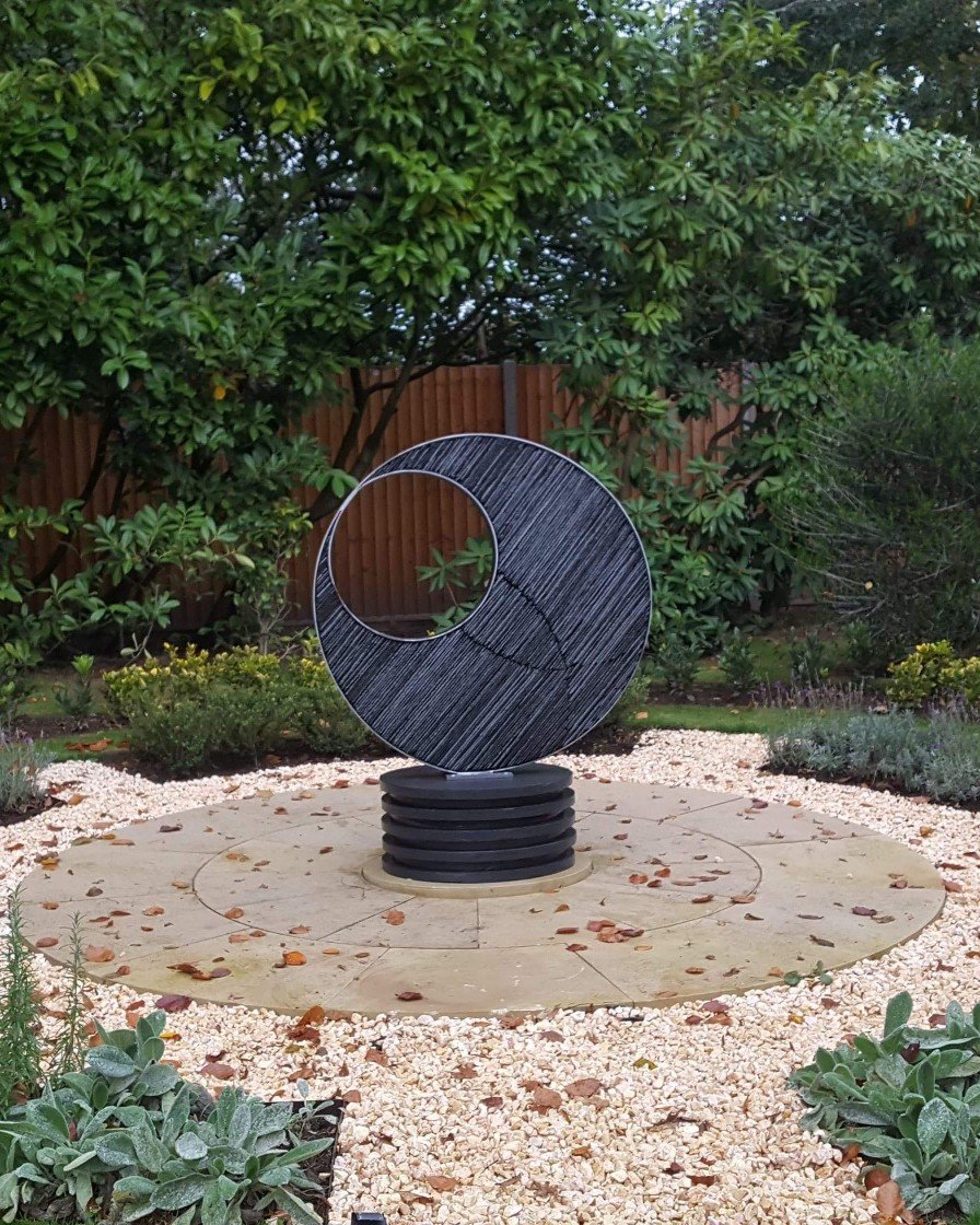Metamorphic Eclipse: Steel and Slate Symphony. Witness the convergence of form and eternity in this mesmerizing sculpture. Two steel circles intertwine, one filled with stacked slate, hinting at the inception of a double helix, while the empty circle