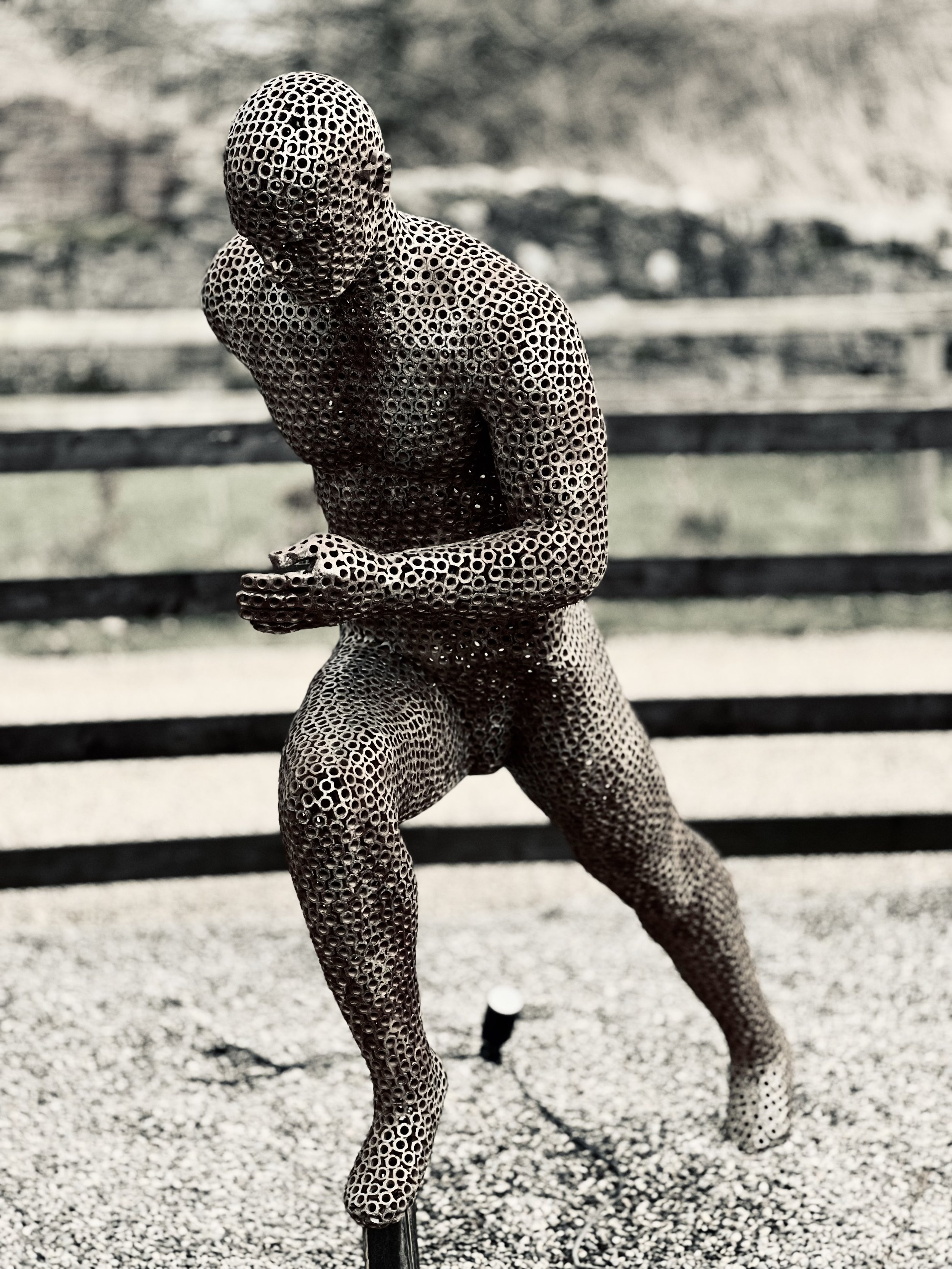 A metal sculpture of a running man, designed by Shaun Gagg in a dynamic pose with one foot lifted off the ground. The figure is made of nuts, creating a semi-transparent, honeycomb-like structure.