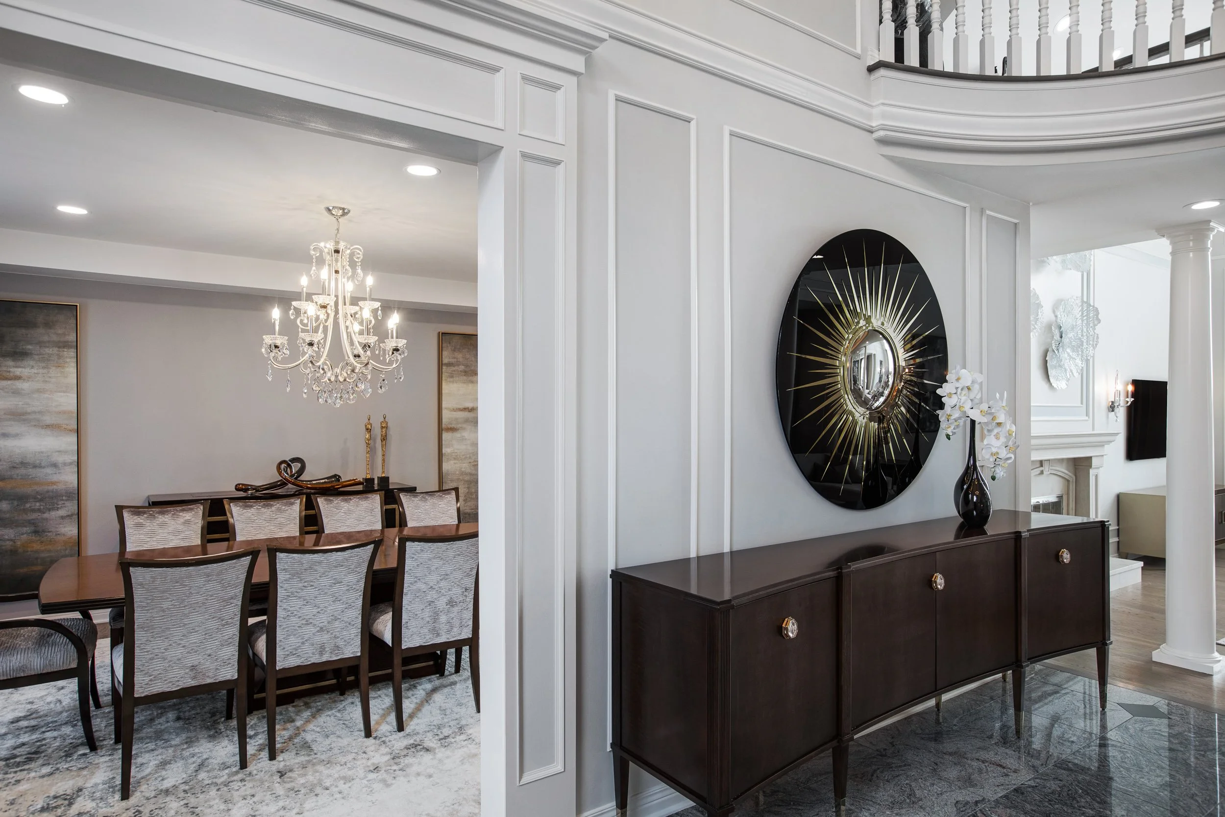 Elegant dining room with a large wooden table surrounded by chairs, a chandelier overhead, abstract wall art, and a decorative wall feature with a sunburst mirror above a console table.