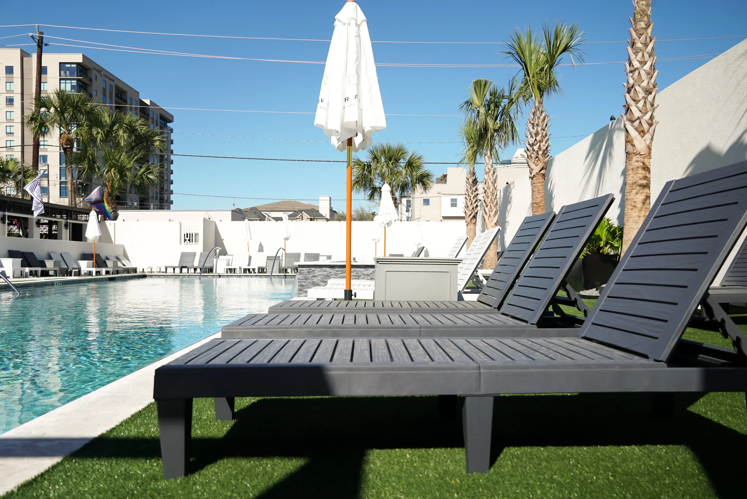 Empty swimming pool area with lounge chairs, white umbrellas, and palm trees under a clear blue sky in an urban setting.