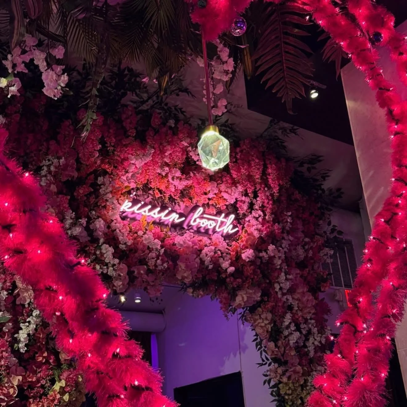 A decorated wall with pink and red flowers, pink feather boas with lights, a neon sign saying 'kissing booth,' and a geometric glass hanging light fixture.