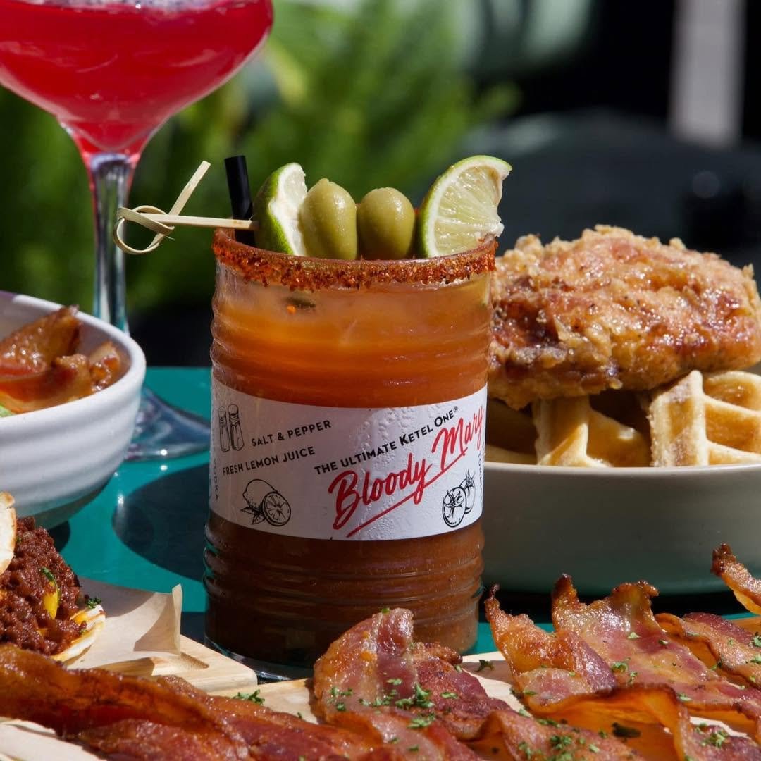 A Bloody Mary cocktail garnished with lime wedges and green olives, surrounded by fried chicken, waffle, bacon, and other dishes on a green table.