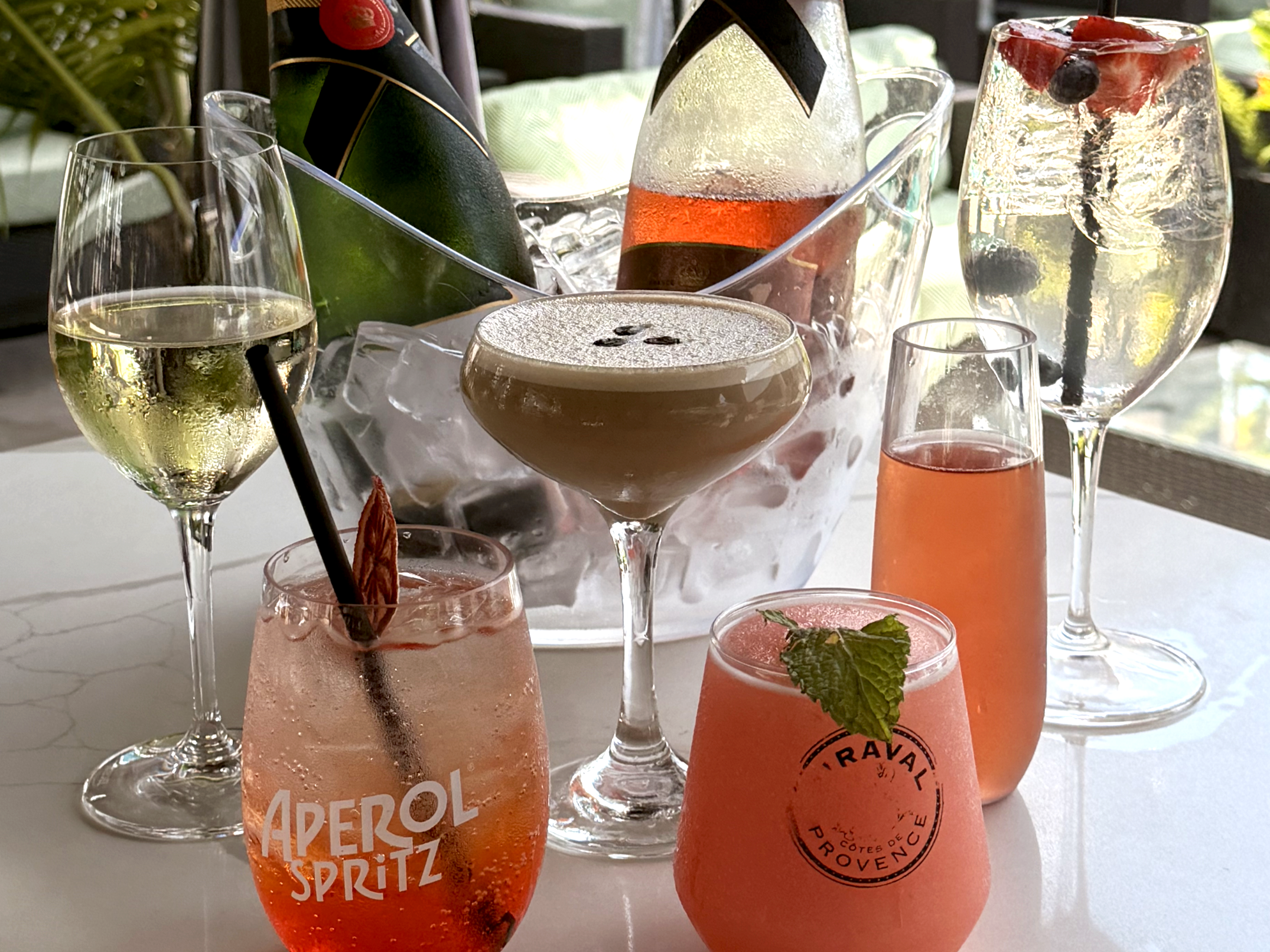 A table with various cocktails in different glasses, some with fruit garnishes, and bottles of sparkling beverages in a cooler, outdoors with natural light.