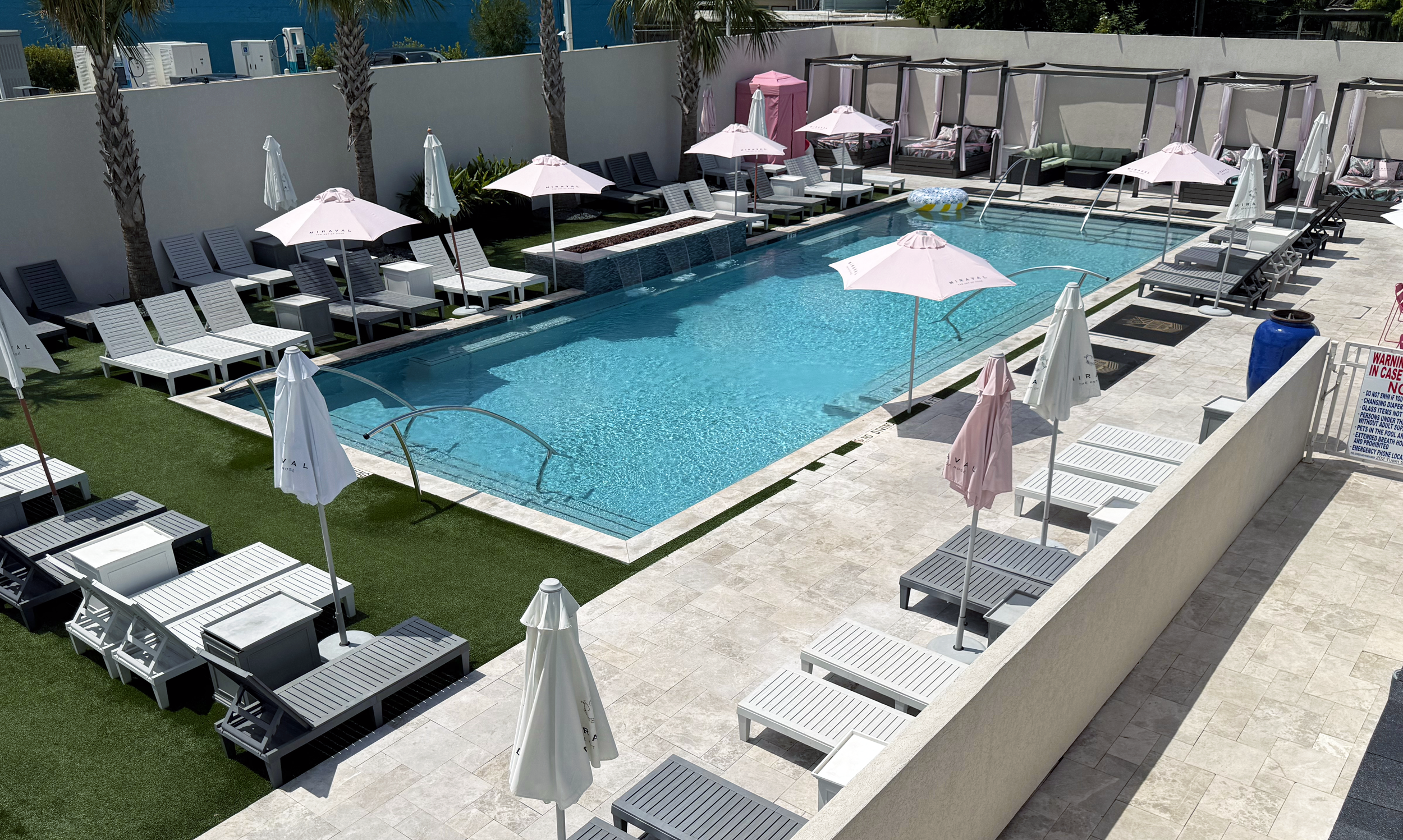 An outdoor swimming pool area with lounge chairs, umbrellas, and cabanas. There are palm trees along the perimeter and a white privacy wall surrounding the area.