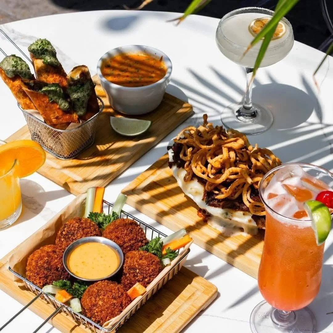 A table with various dishes including fried appetizers with dipping sauce, a bowl of soup, a large sandwich with fried onions on top, and drinks such as an orange fruit drink, a clear cocktail with citrus slices, and a pink cocktail with fruit garnis