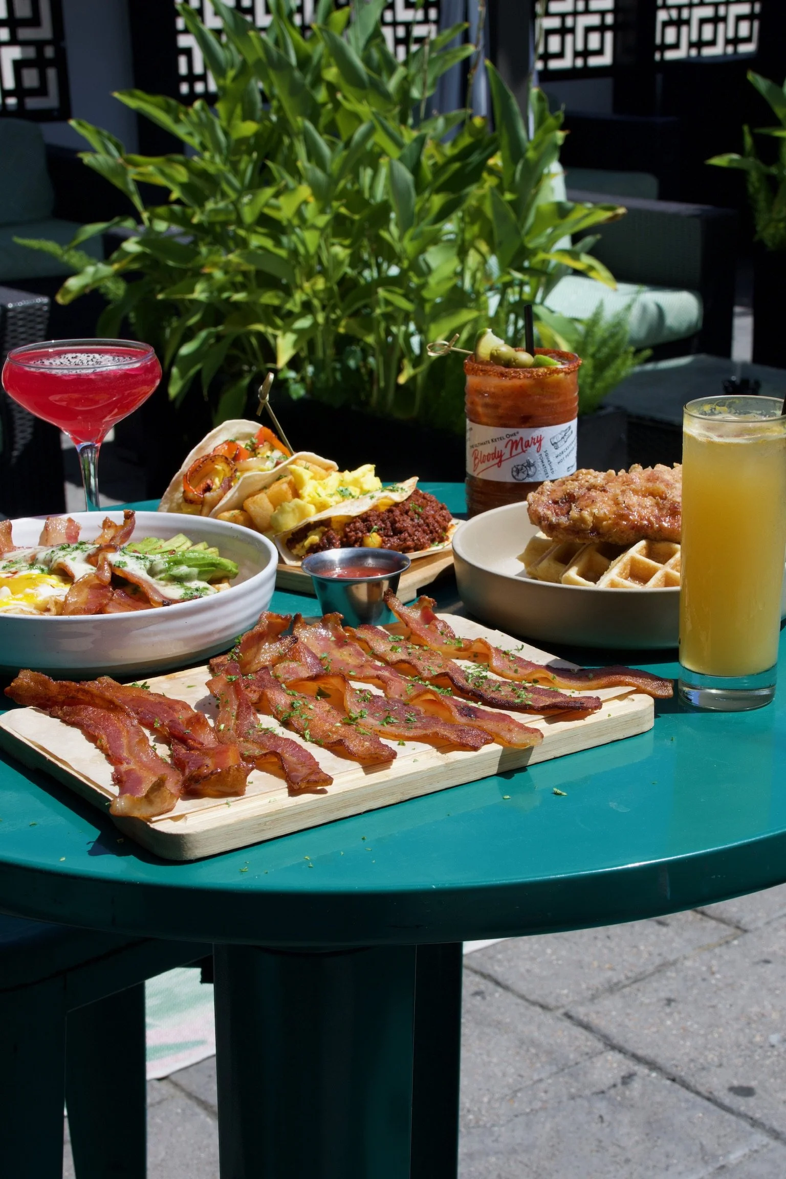 Outdoor table with various foods including bacon, waffles, pizza, a sandwich, a taco, and drinks, with greenery and patio furniture in the background.